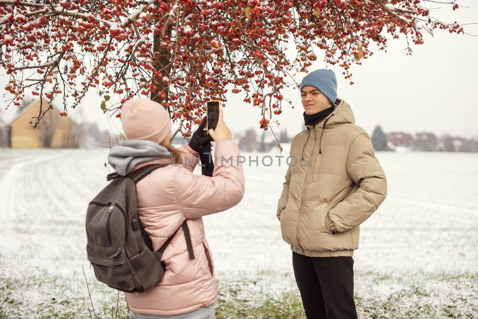 Winter Romance: Girl in Pink Winter Jacket Photographing Boy Against Snow-Covered Red Tree and Field. Embrace the winter magic in this enchanting image, where a girl in a pink winter jacket captures a moment as she photographs her companion against the backdrop of a snow-covered red tree and field. The photograph beautifully conveys the essence of winter romance and the serene beauty of a snowy landscape.