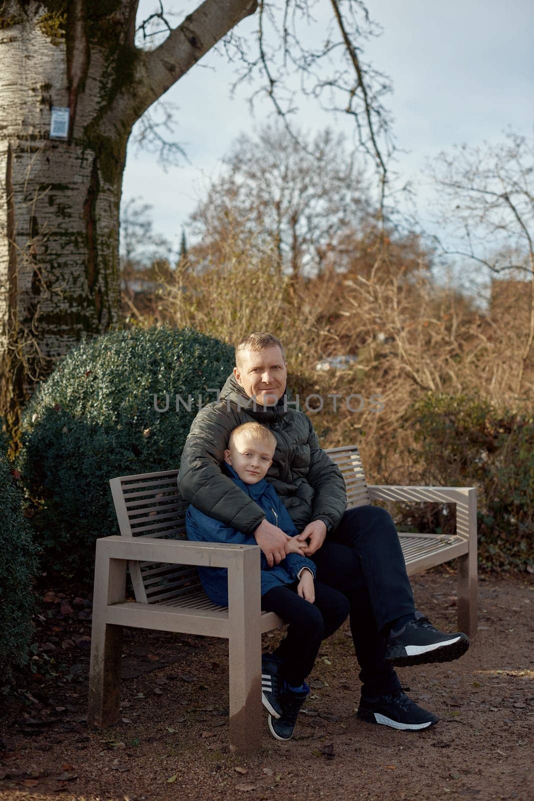 Autumnal Family Affection: Father, 40 Years Old, and Son - Beautiful 8-Year-Old Boy, Seated in the Park. by Andrii_Ko