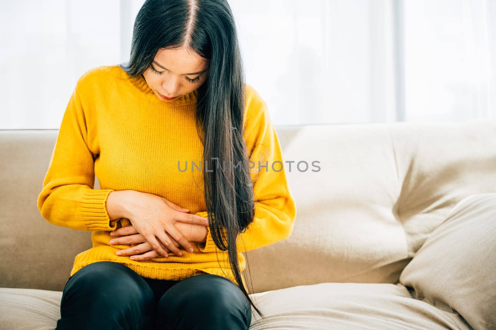 Asian woman seated on a home couch experiences abdominal pain by Sorapop