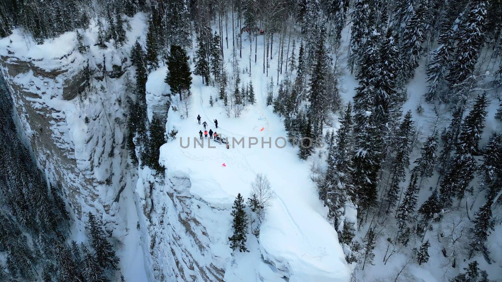 Winter mountain cliff covered by snow, ice, and fur trees. Clip. Stunning frozen winter nature, aerial view of tourists enjoying the day on the cliff edge. by Mediawhalestock