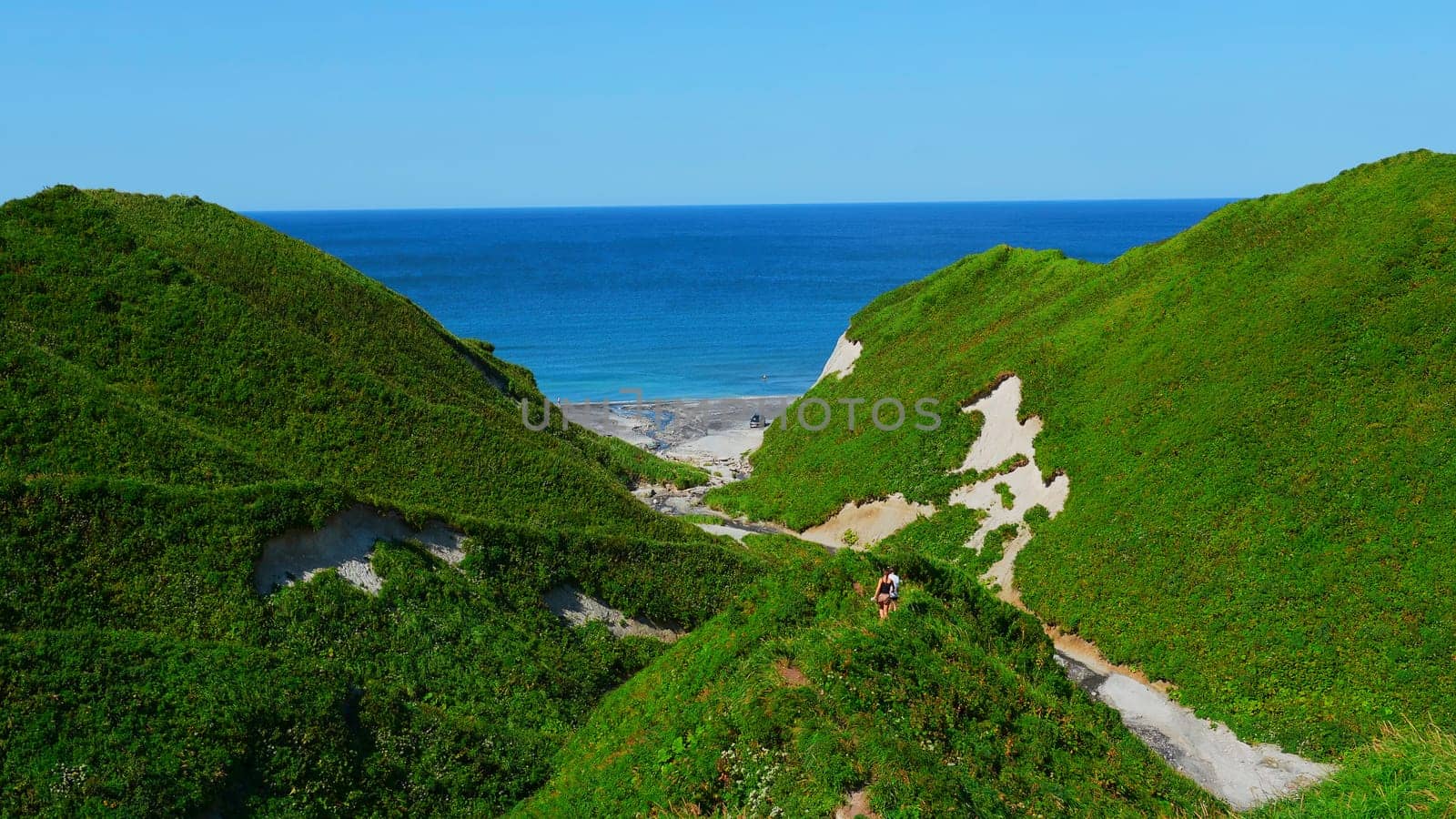 Couple walking along path on hills on background of sea. Clip. Top view of hills with trails and green grass on background of sea coast. Beautiful summer hills by sea on sunny day.