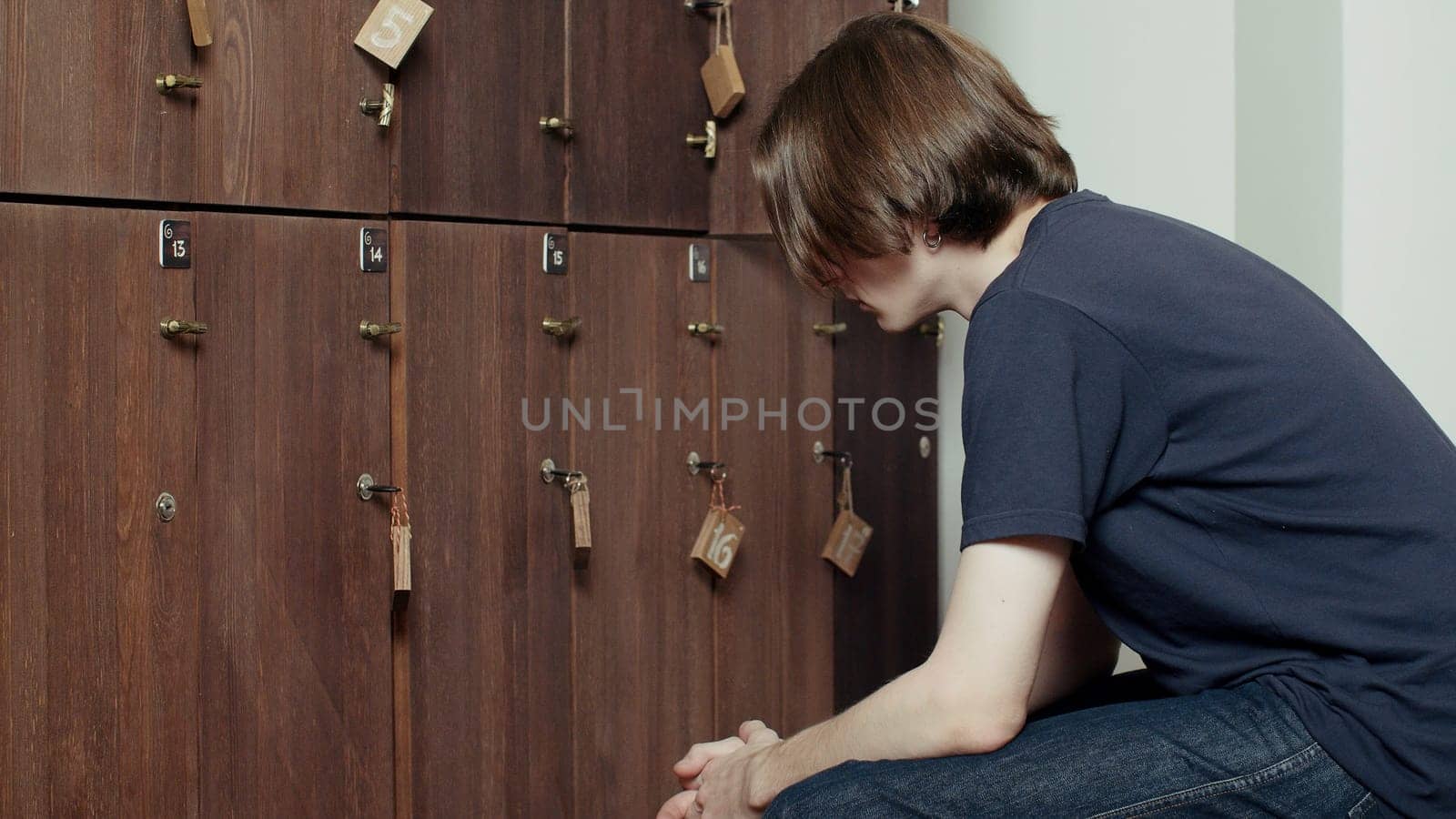 Young tired man sitting against a wooden gym cabinets. Media. Locker room in the gym. by Mediawhalestock