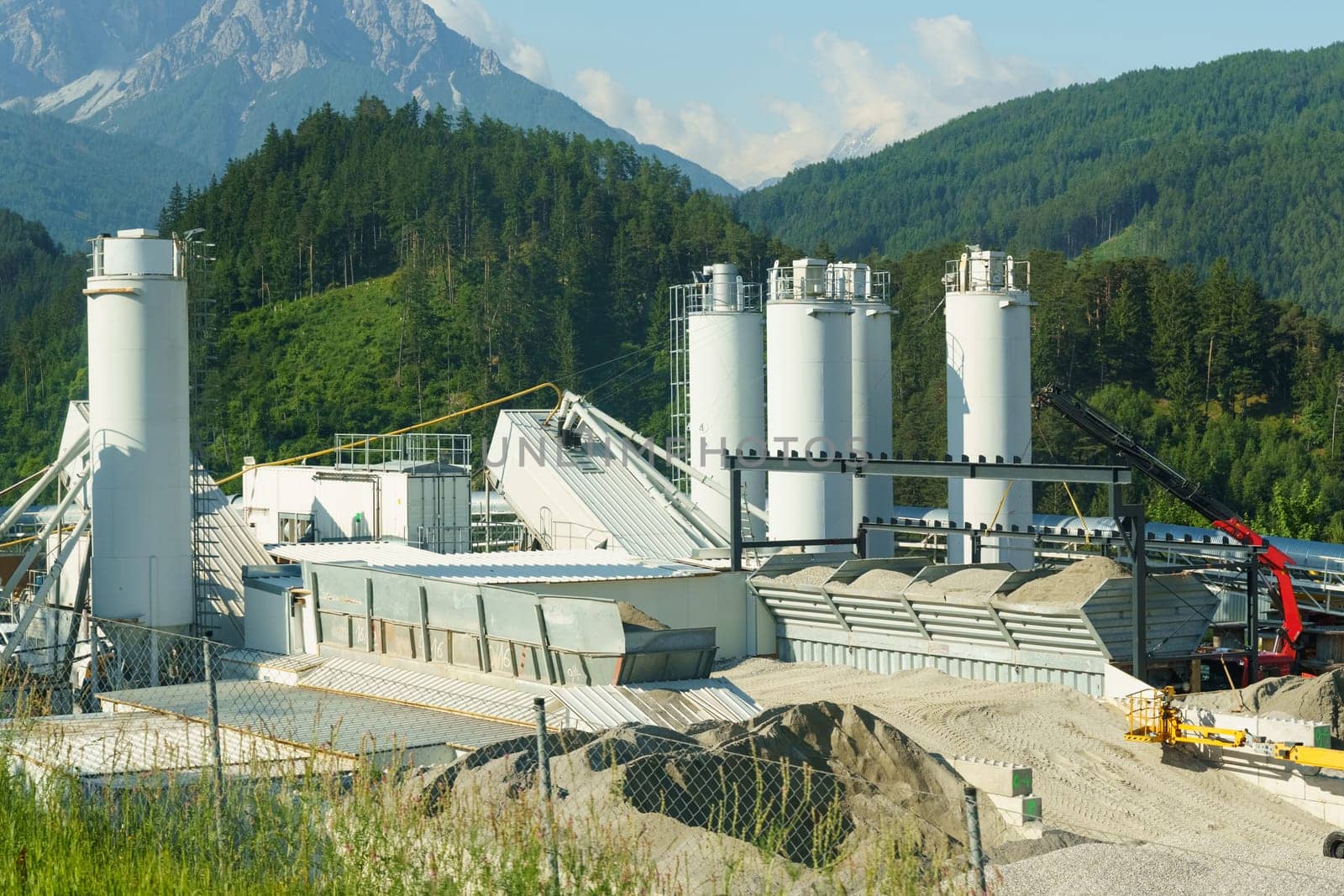 A busy cement factory with towering silos set against a backdrop of lush mountains, conveying large-scale industrial activity in a natural setting.