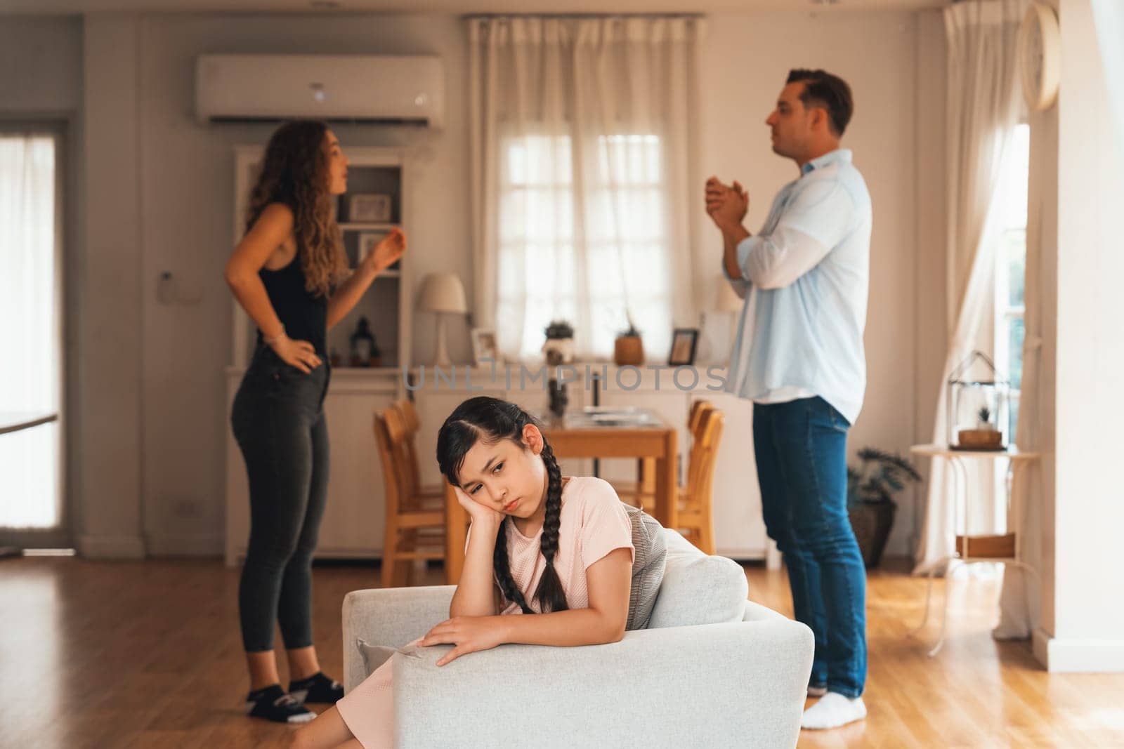 Annoyed and unhappy young girl sitting on sofa trapped in middle of tension by her parent argument in living room. Unhealthy domestic lifestyle and traumatic childhood develop to depression.Synchronos