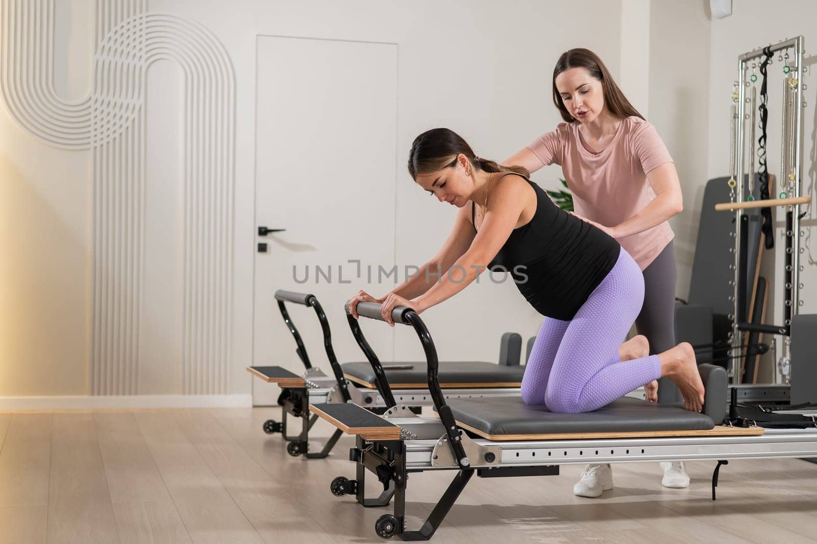 A pregnant woman works out on a reformer exercise machine with a personal trainer