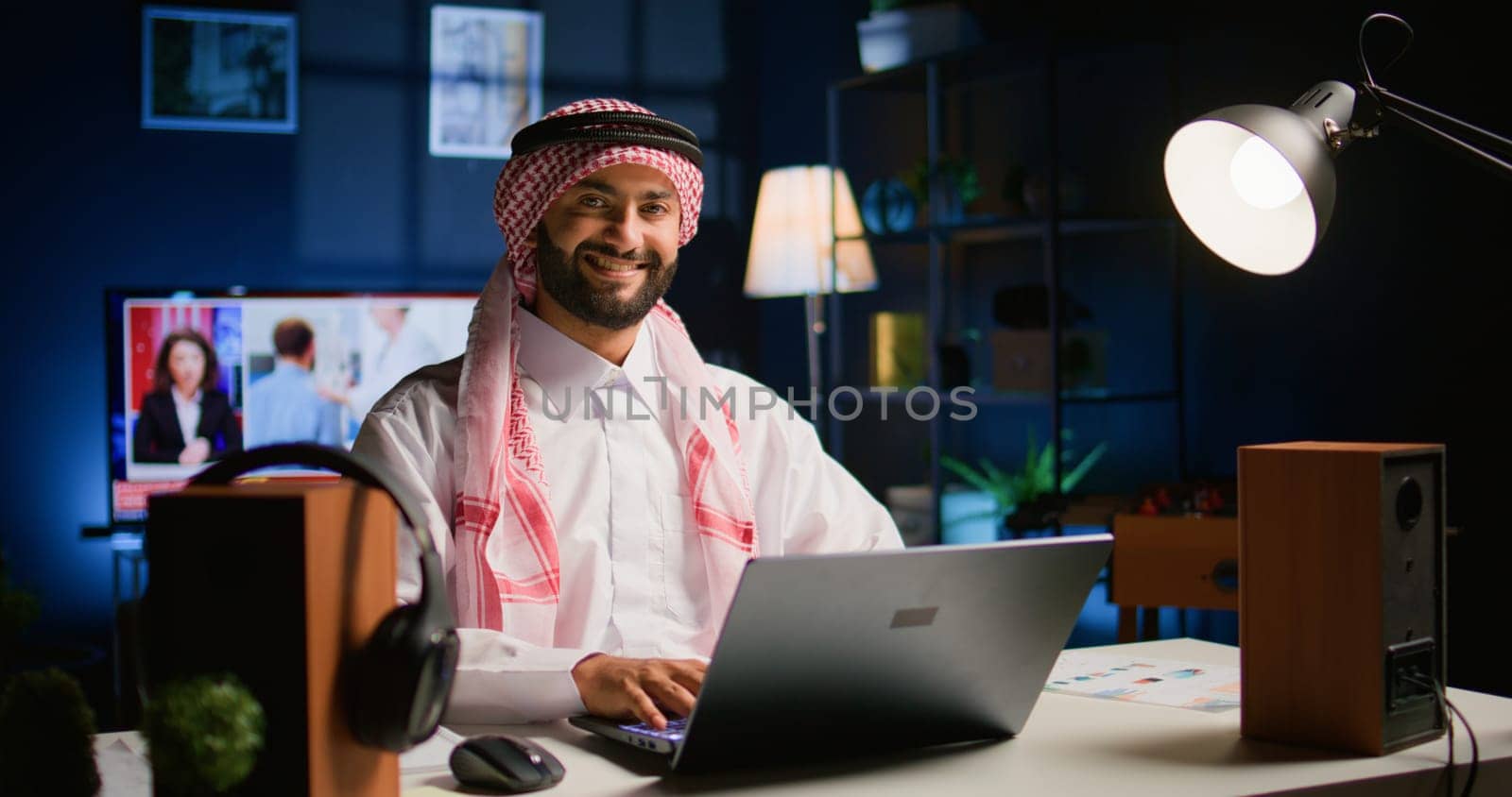 Happy arabic teleworker answering job emails in stylish apartment. Cheerful muslim employee remotely working, typing data on laptop with opened tv as background noise, handheld camera shot