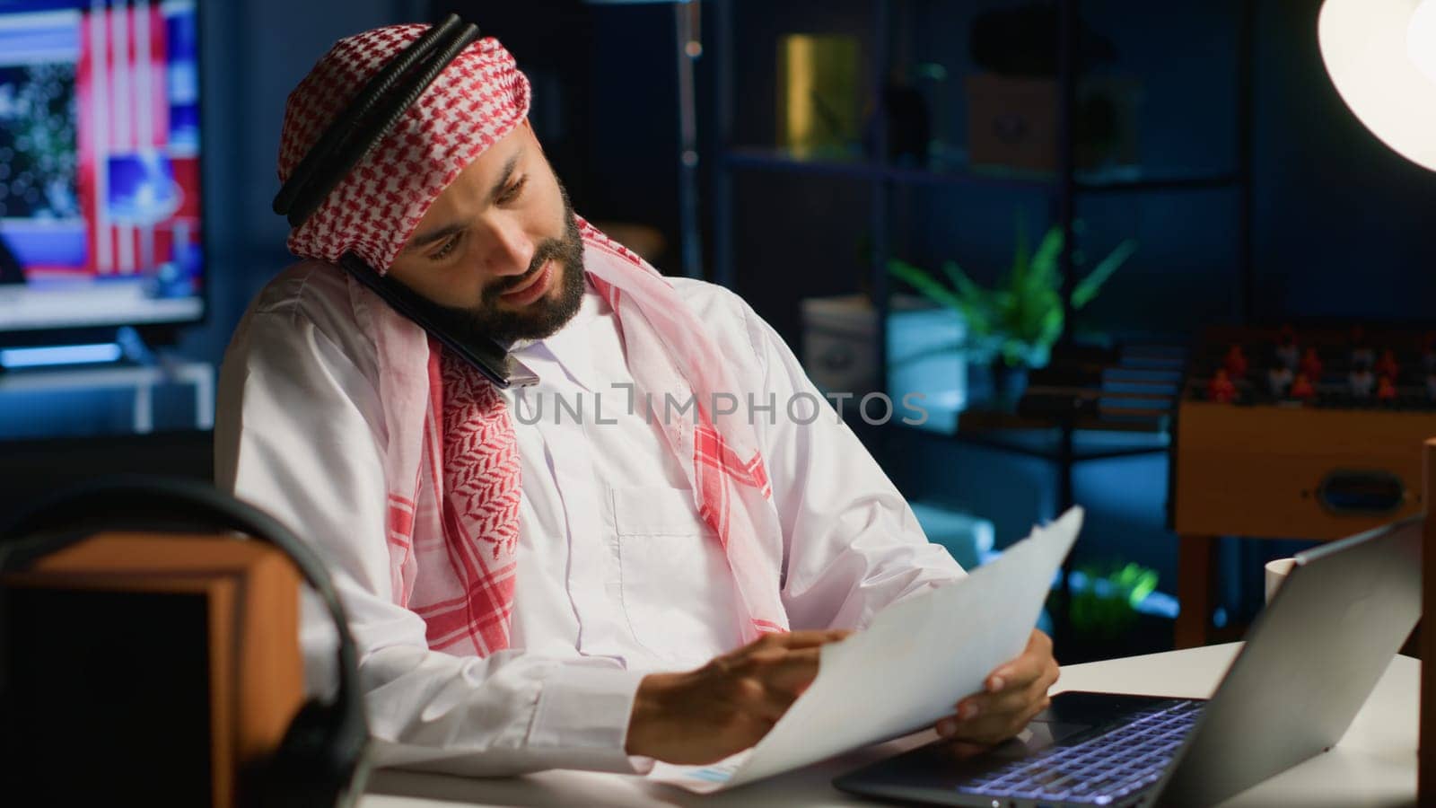 Arabic employee having discussion with coworker in phone call while remotely working from cozy home office. Teleworker talking with colleague using smartphone, discussing paperwork