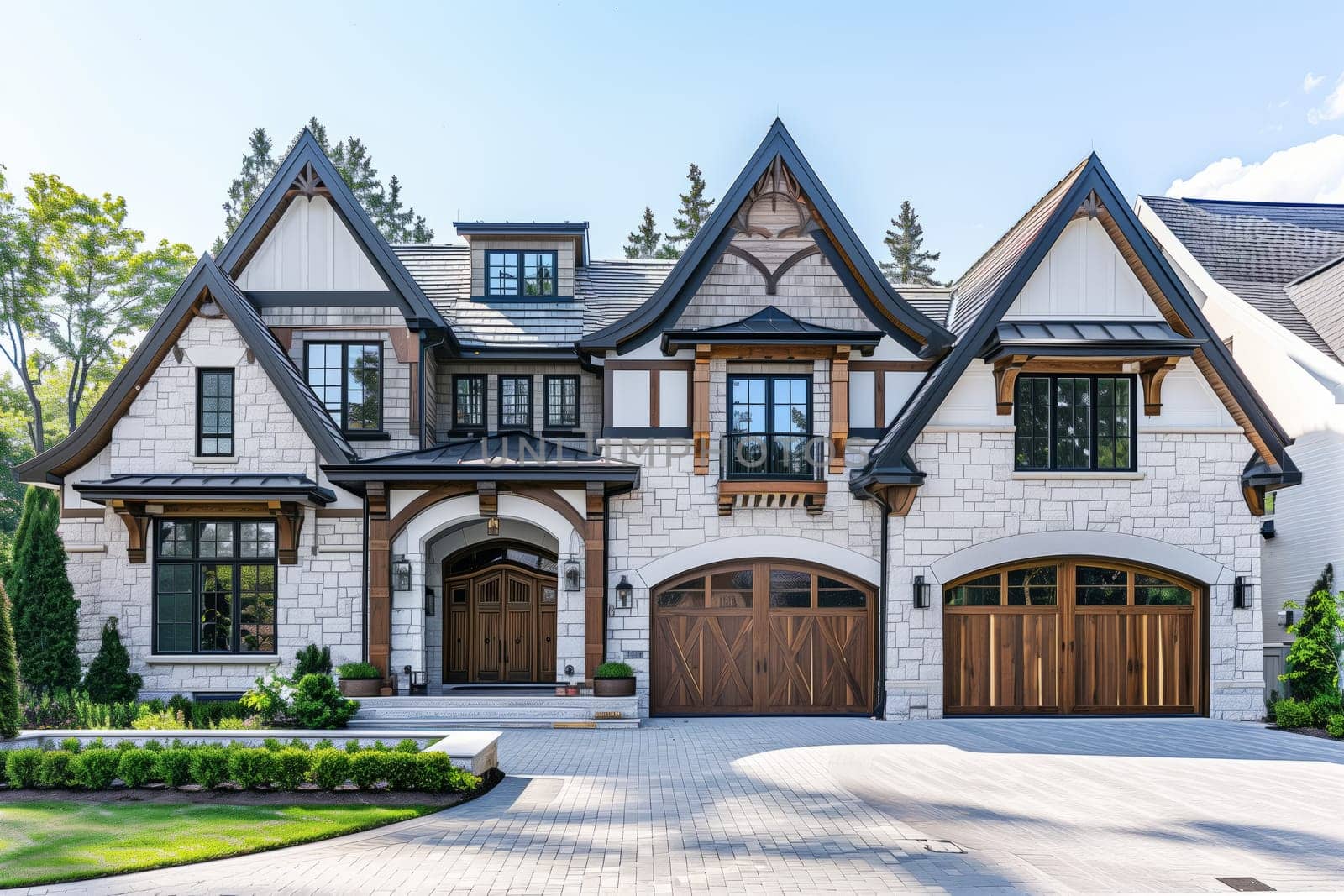 A white house with black trim and wooden garage doors stands tall amidst green grass and lush trees, under a clear blue sky