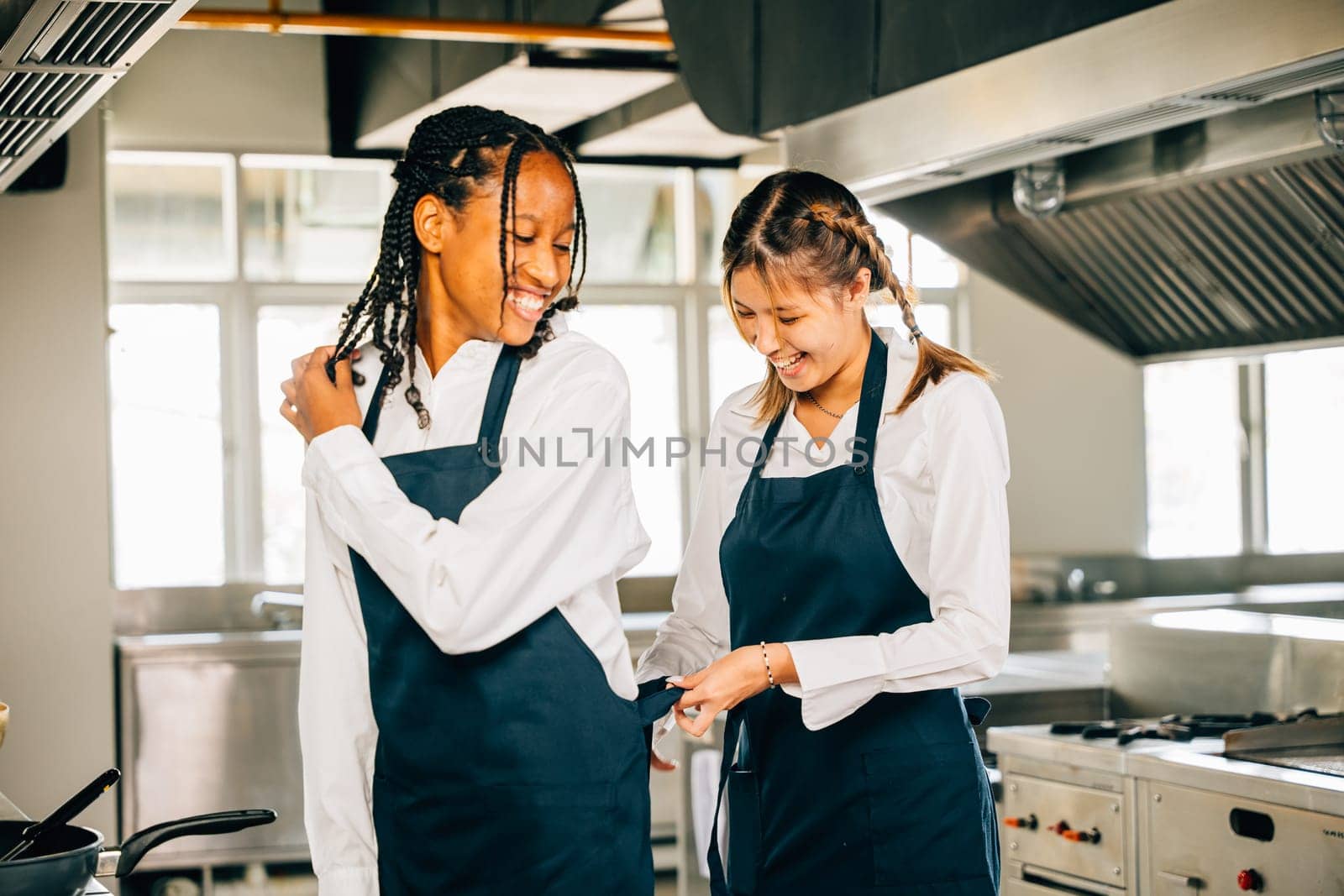 Girlfriend chef aids woman friend tie apron in a restaurant kitchen. Two adults in uniform prepare for food education and service in a professional setting.
