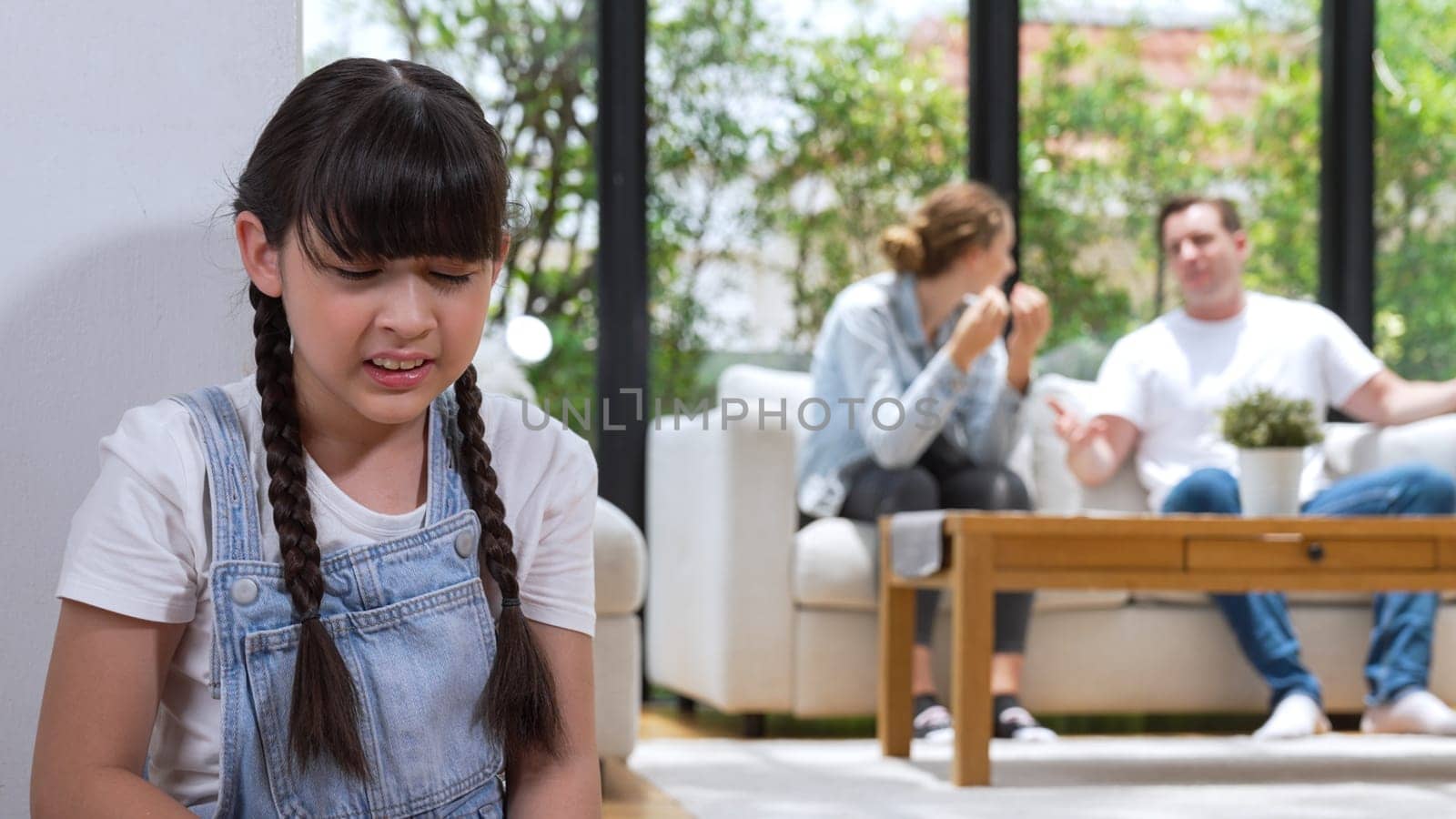 Stressed and unhappy young girl huddle in corner, cover her ears blocking sound of her parent arguing in background. Domestic violence at home and traumatic childhood develop to depression. Synchronos