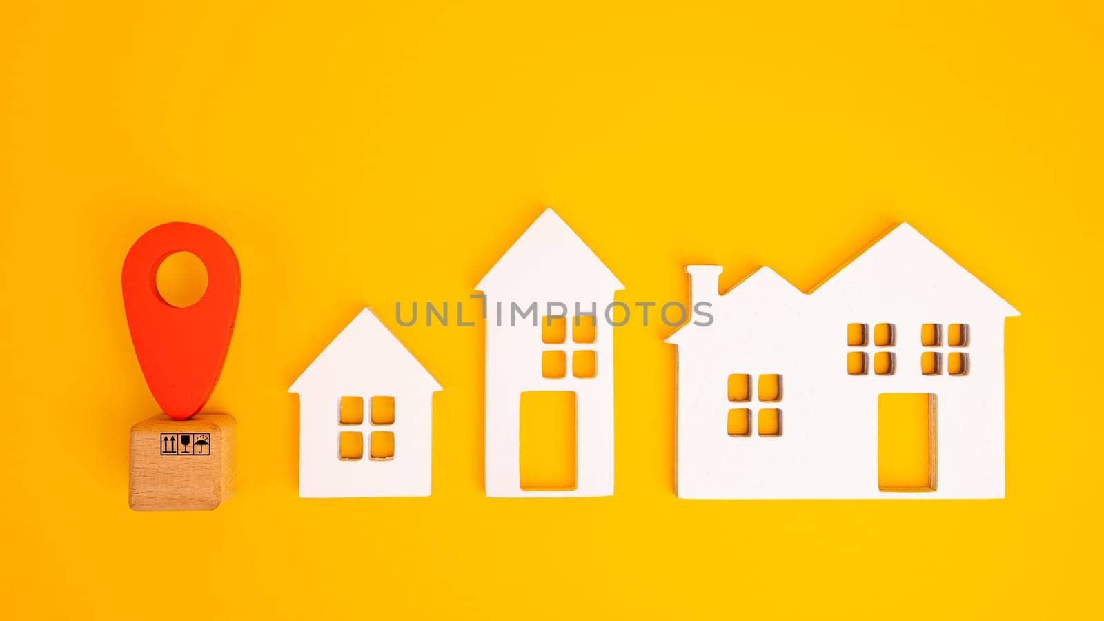 Cardboard boxes and a red position pin in city of white toy house on yellow background. Locating packages and goods.