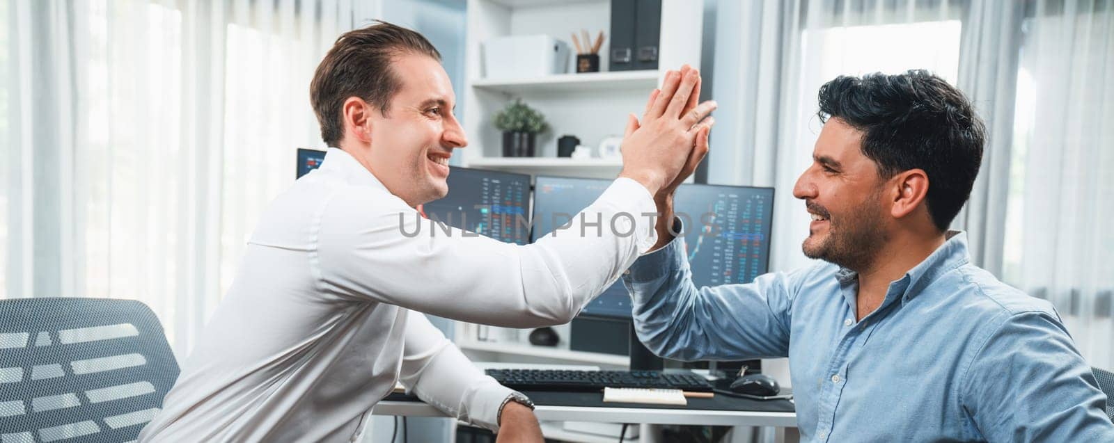Cheerful two smiling stock traders giving high five on panorama view to success of digital technology in dynamic data monitor background. Achievement investors in business at workplace. Sellable.