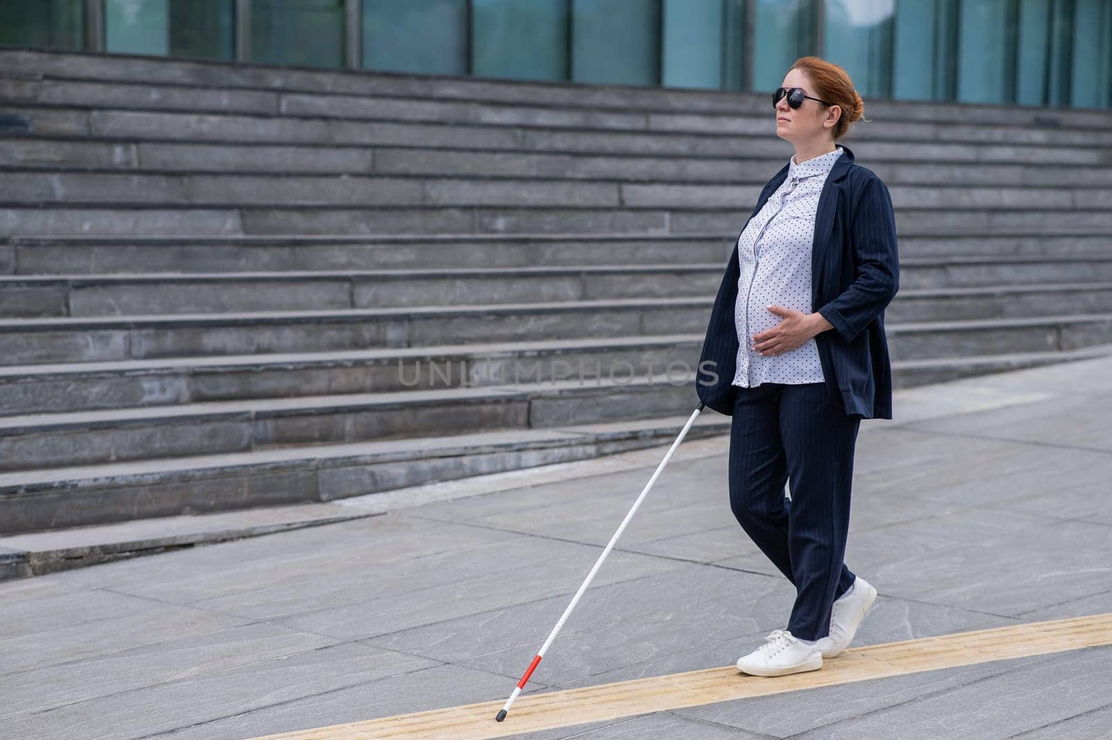 Blind pregnant businesswoman walking along tactile tiles with a cane. by mrwed54