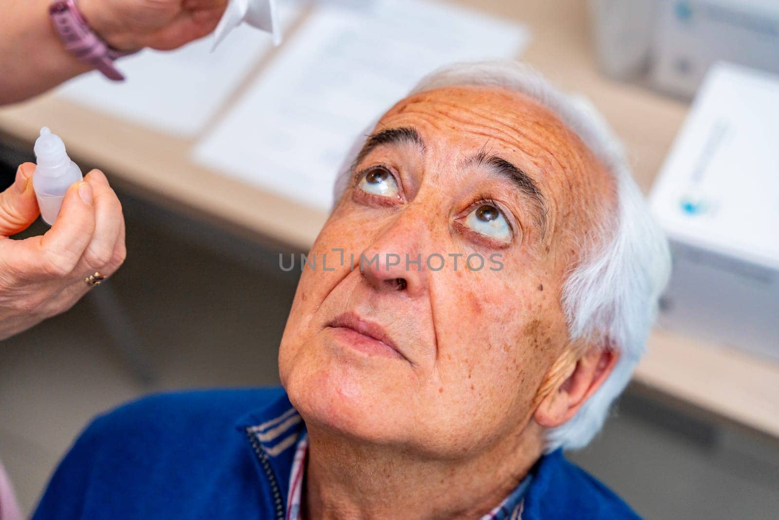 Close-up top view of a senior man looking up while ophthalmologist applying drops in the eyes in a clinic