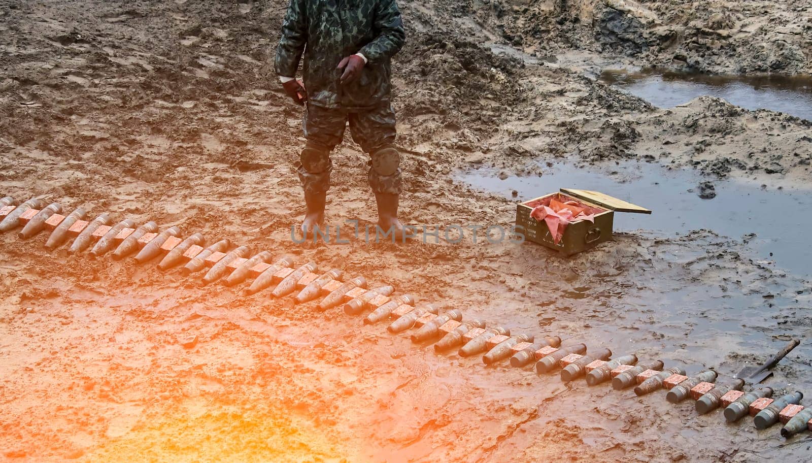 A soldier in uniform prepares weapons in a muddy field, surrounded by ammunition boxes. The low angle shot makes the soldier look powerful and intimidating, capturing the horror and futility of war.