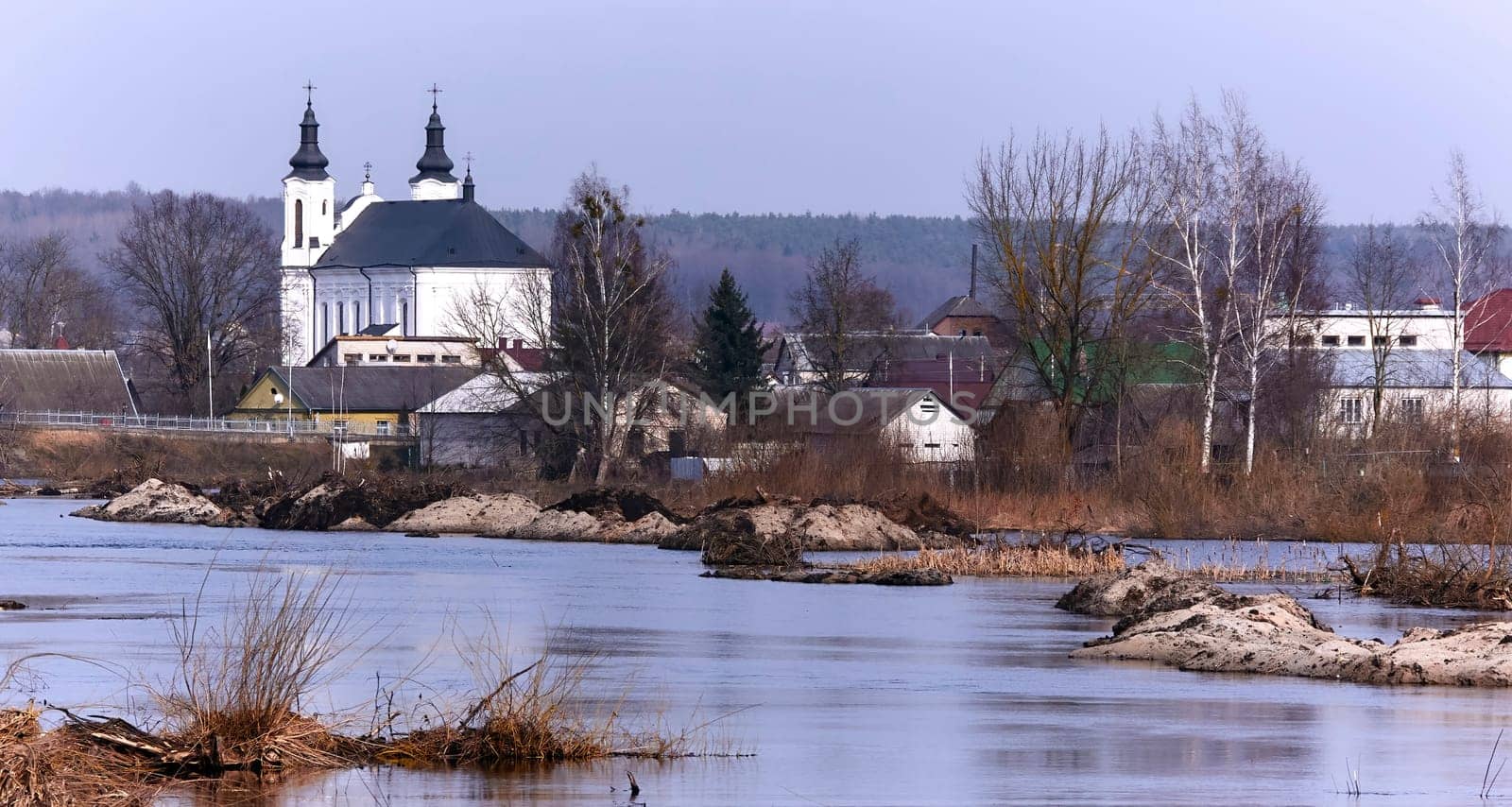 A picturesque European village by a serene river, surrounded by greenery and colorful flowers. White church, quaint houses, blue sky with fluffy clouds - a perfect setting for a peaceful spring day.