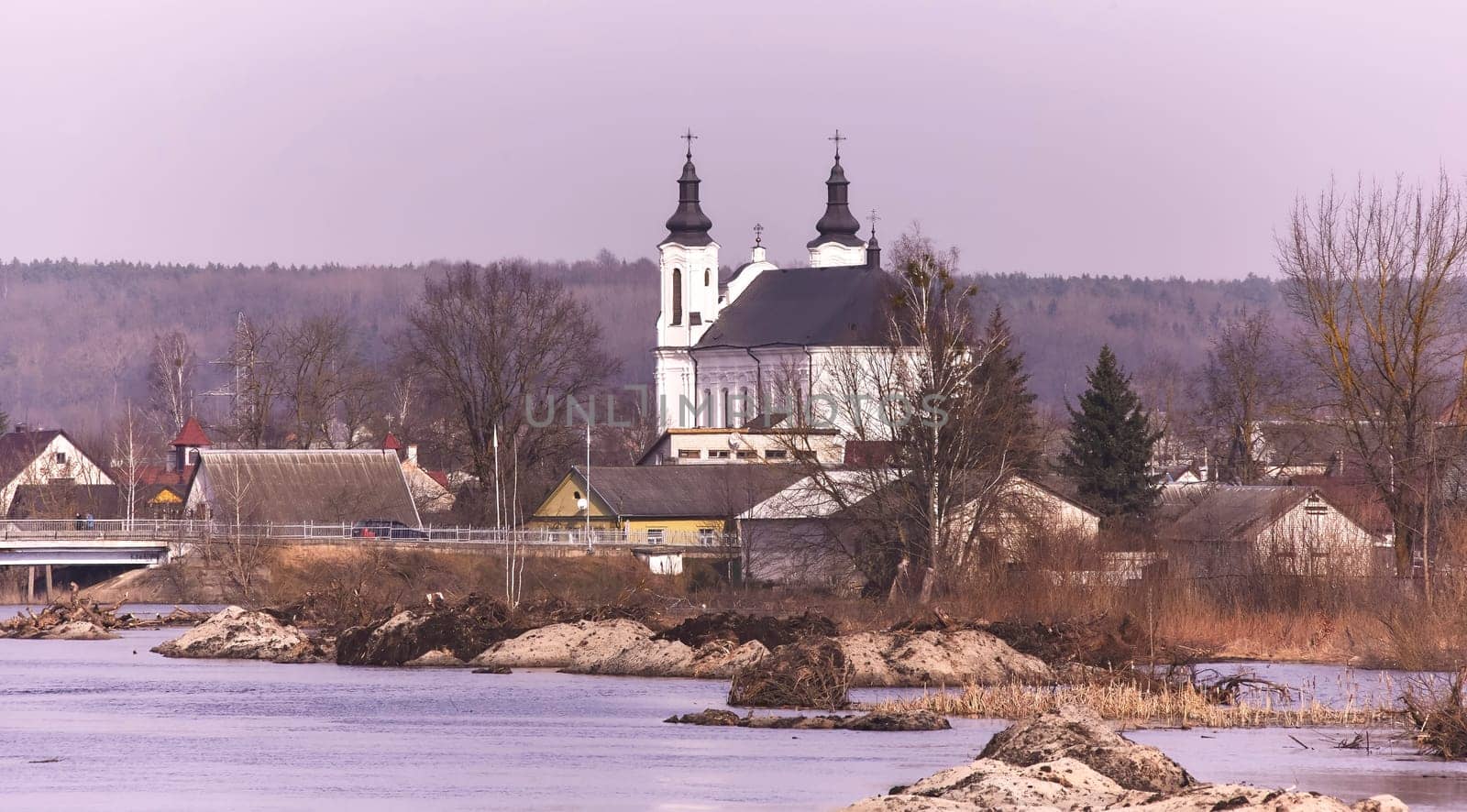 A picturesque European village by a serene river, surrounded by greenery and colorful flowers. White church, quaint houses, blue sky with fluffy clouds - a perfect setting for a peaceful spring day.