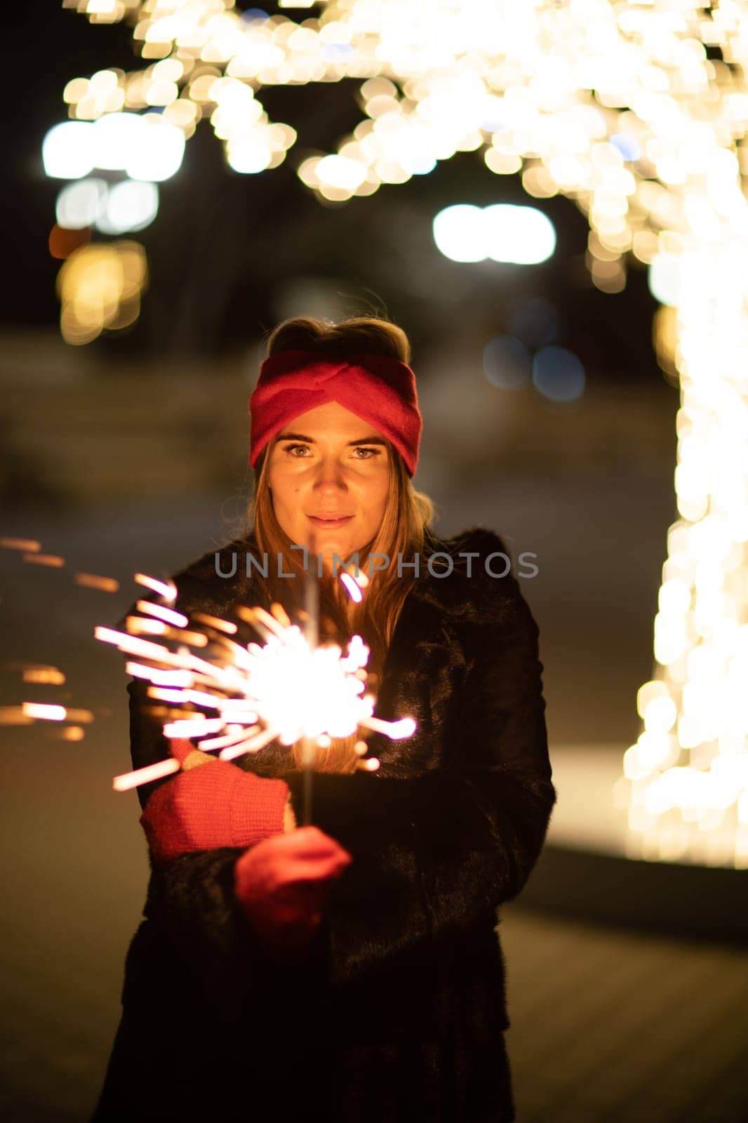 Woman holding sparkler night while celebrating Christmas outside. Dressed in a fur coat and a red headband. Blurred christmas decorations in the background. Selective focus.