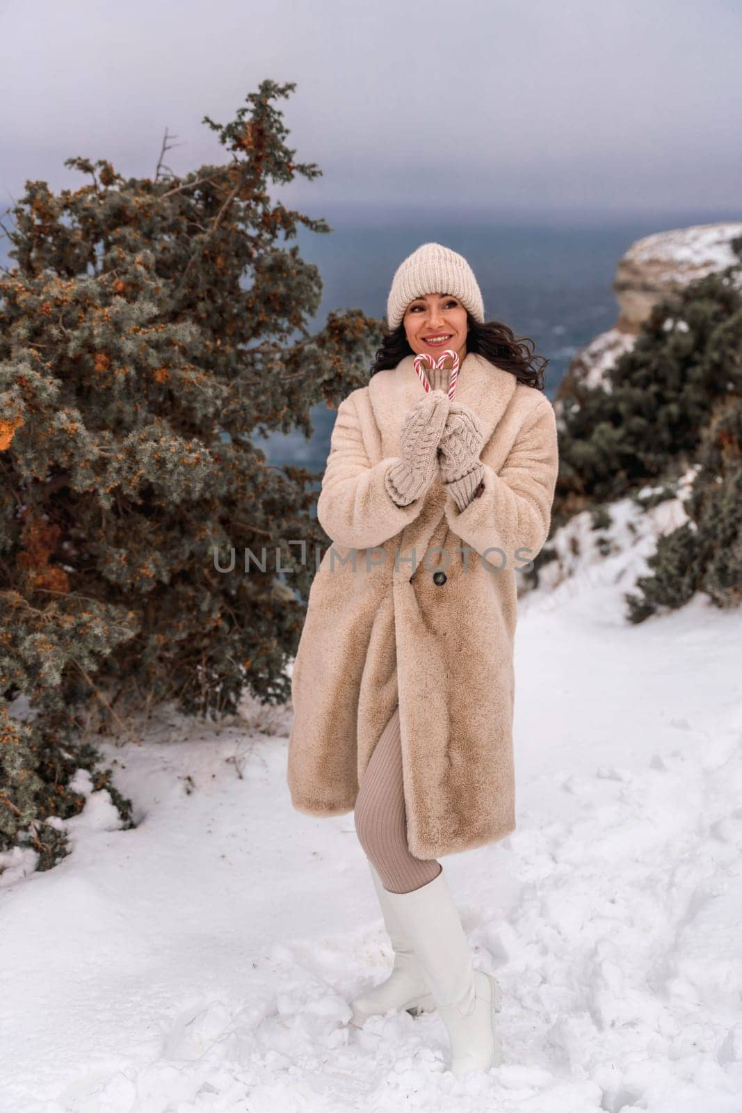 Woman snow sea. Smiling woman in knitted hat, mittens and beige coat holding lollipops candy canes in her hands in shape of heart against the backdrop of the sea. by Matiunina