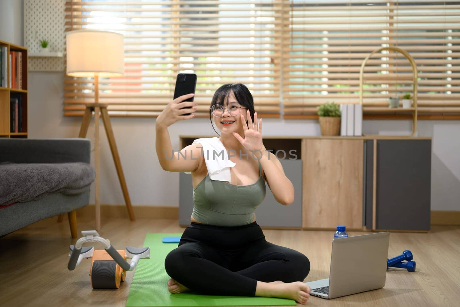 Pretty young woman sitting on exercise mat and making video call over mobile phone at home.