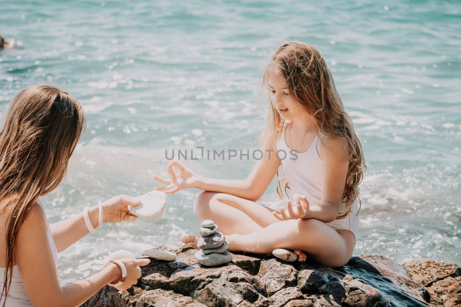 Woman with daughter bilds stones pyramid on seashore on a sunny day on the blue sea background. Happy family holidays. Pebble beach, calm sea. Concept of happy vacation on the sea, meditation, spa by panophotograph