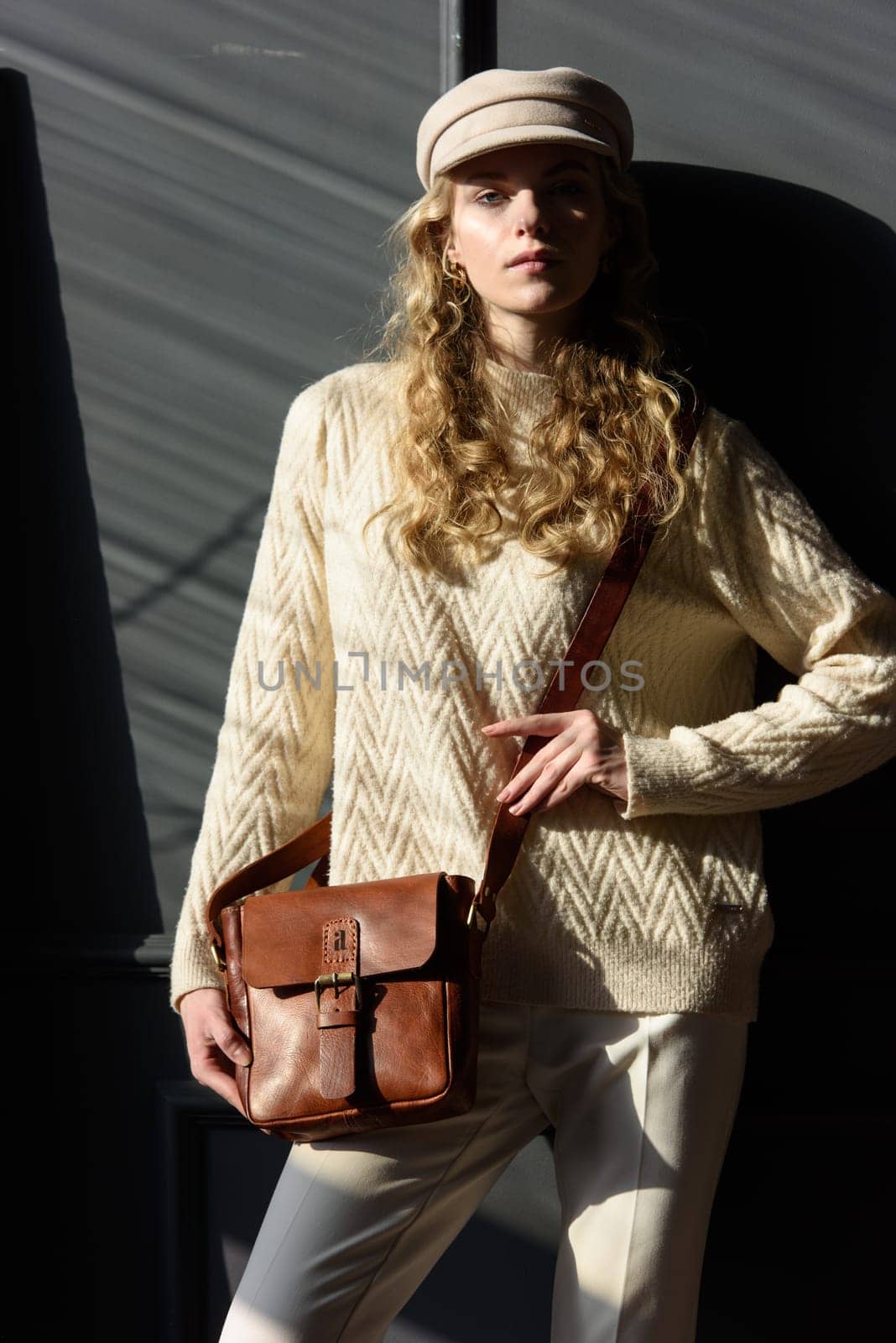 Studio portrait of beautiful woman with a curly blond hair holding brown bag, posing on gray background. Model wearing stylish cap, sweater and classic trousers