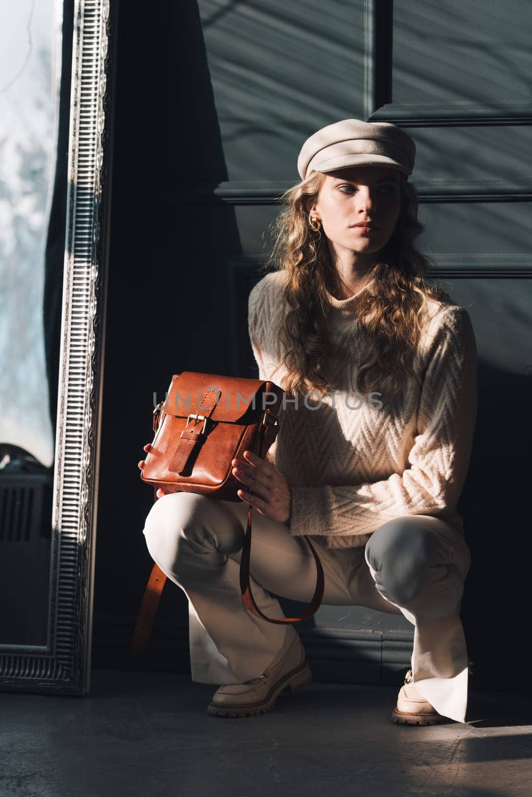 Studio portrait of beautiful woman with a curly blond hair holding brown bag, posing on gray background. Model wearing stylish cap, sweater and classic trousers