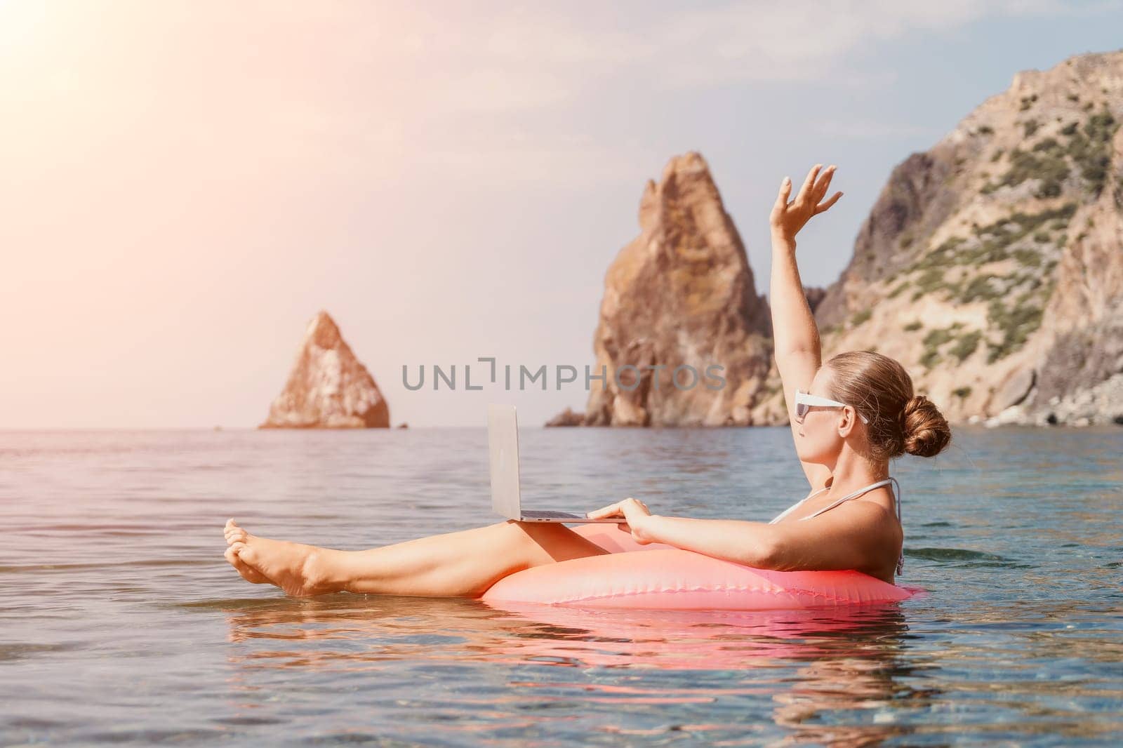 Woman freelancer works on laptop swimming in sea on pink inflatable ring. Pretty lady typing on computer while floating in the sea on inflatable donut at sunset. Freelance, remote work on vacation