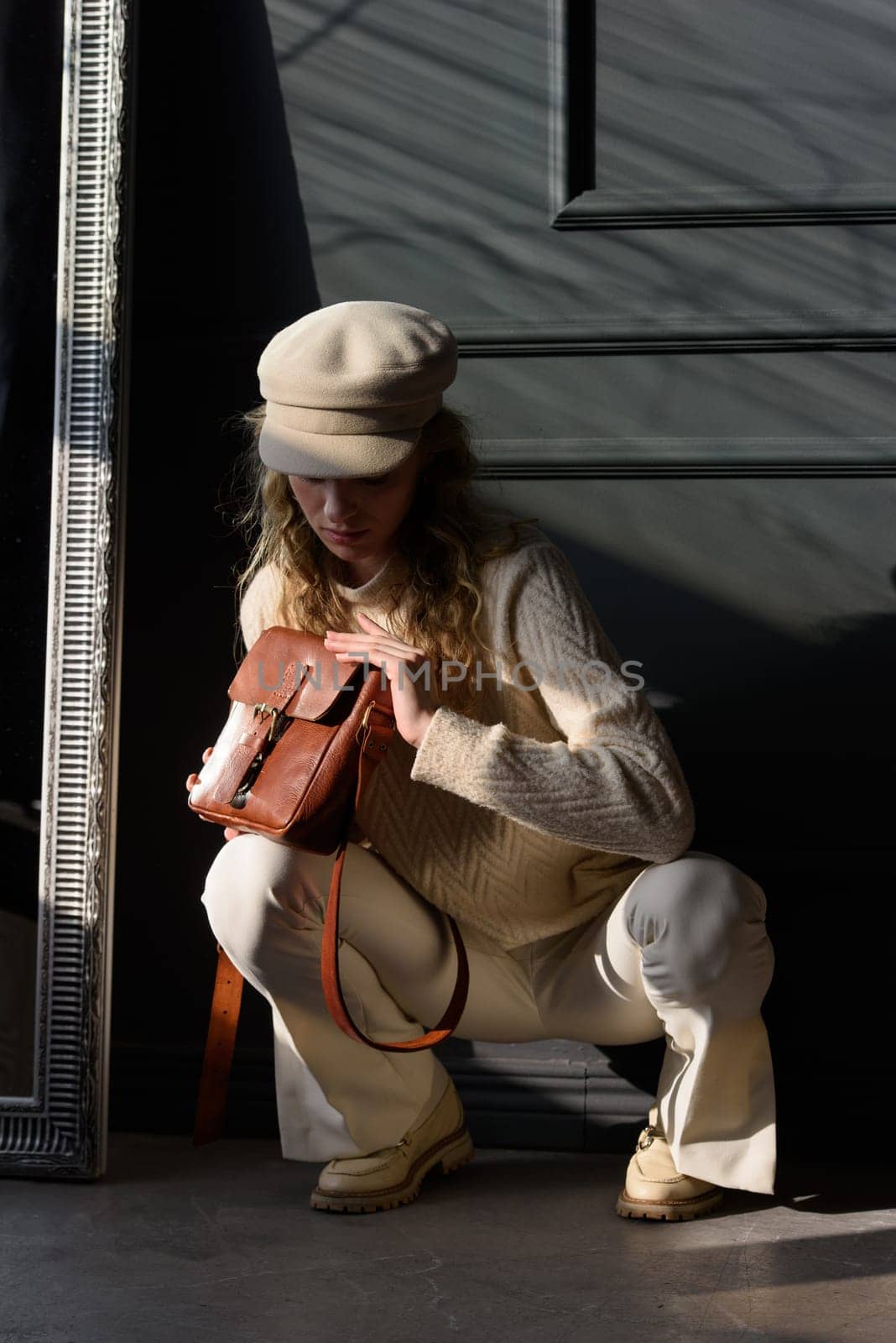 Studio portrait of beautiful woman with a curly blond hair holding brown bag, posing on gray background. Model wearing stylish cap, sweater and classic trousers