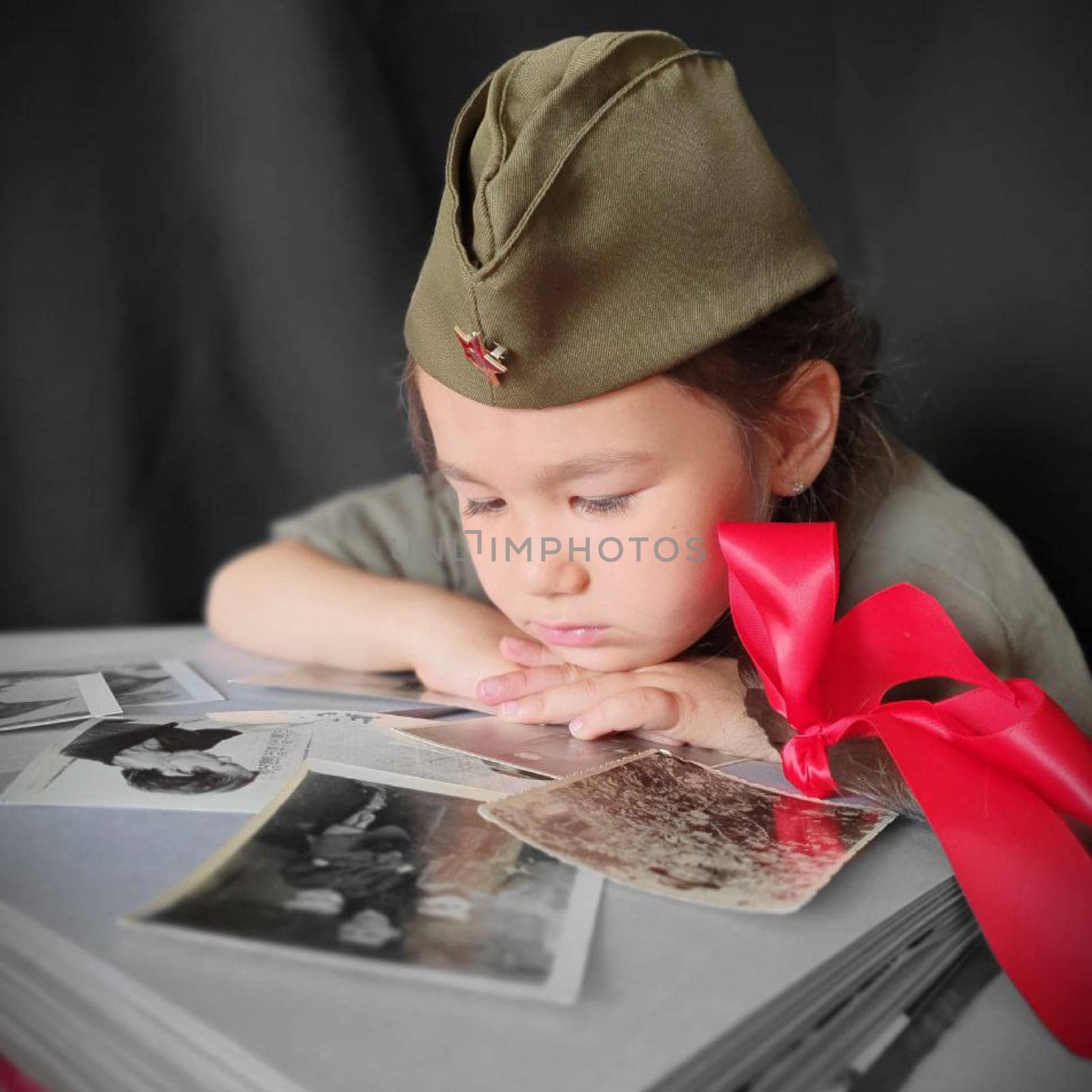 A little girl in a military uniform with two pigtails and red bows is looking at an album with photographs. Portrait photo for Victory Day. 9th May. High quality photo