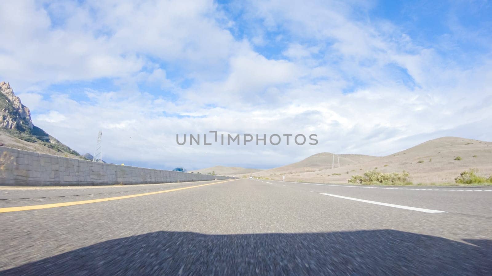 On a crisp winter day, a car cruises along the iconic Highway 101 near San Luis Obispo, California. The surrounding landscape is brownish and subdued, with rolling hills and patches of coastal vegetation flanking the winding road.