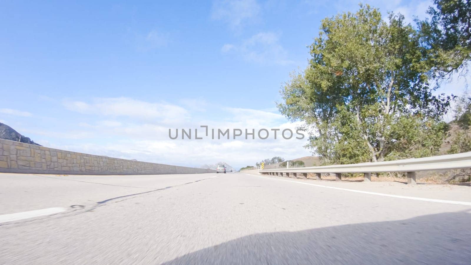 On a crisp winter day, a car cruises along the iconic Highway 101 near San Luis Obispo, California. The surrounding landscape is brownish and subdued, with rolling hills and patches of coastal vegetation flanking the winding road.