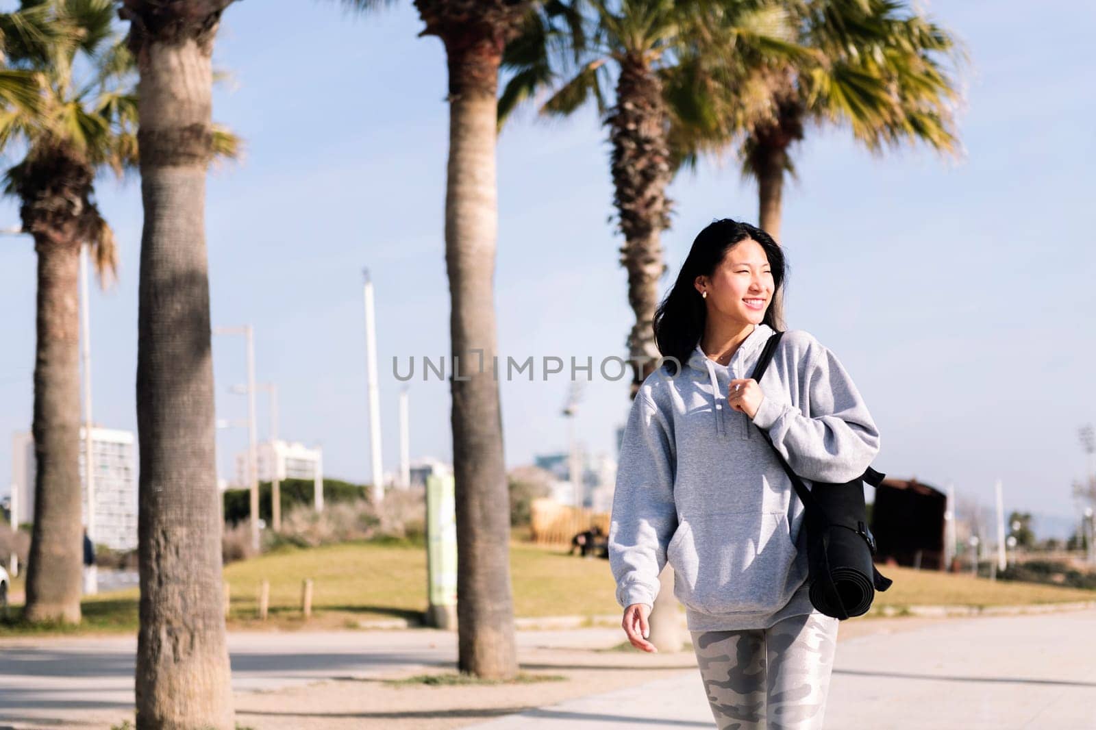 asian woman smiling while walking with yoga mat by raulmelldo