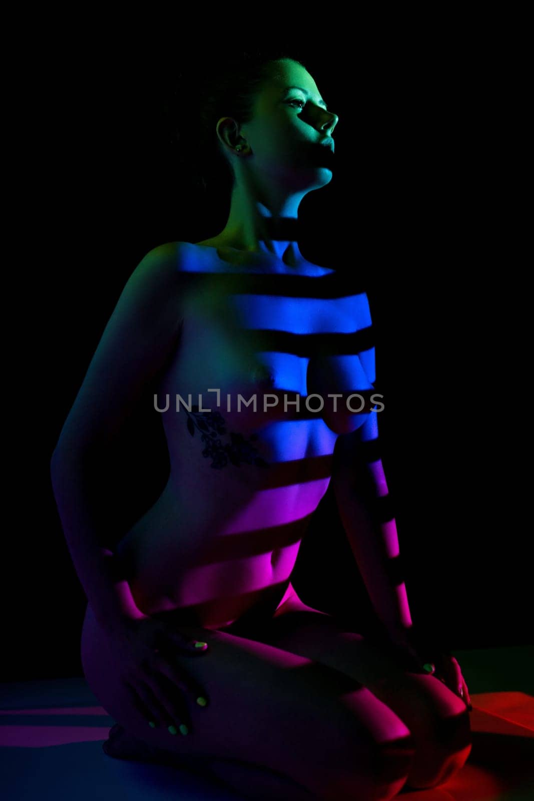 Young seductive nude female with dreamy gaze sitting in multicolored light and looking away in evening on black background