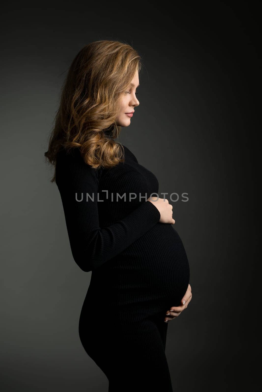 Side view of tranquil pregnant female wearing long black dress and touching tummy while standing on dark background in studio