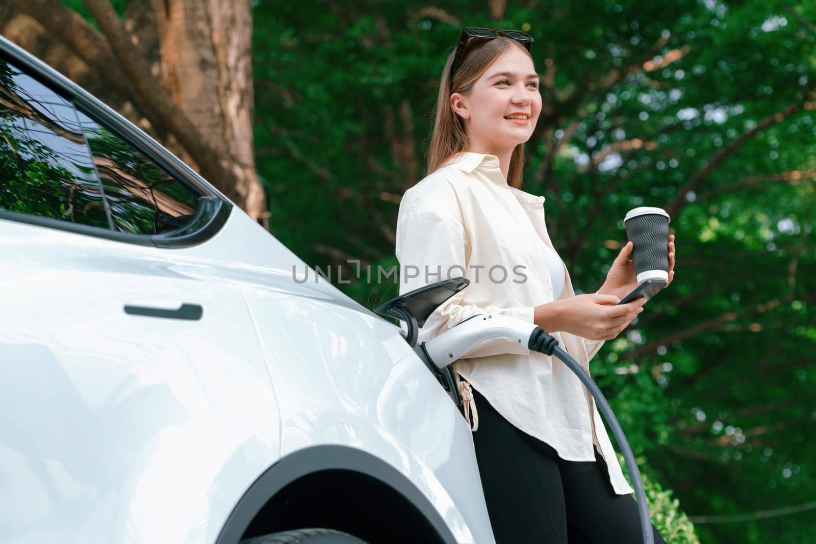 Young woman use smartphone to pay for electricity at public EV car charging station green city park. Modern environmental and sustainable urban lifestyle with EV vehicle. Expedient