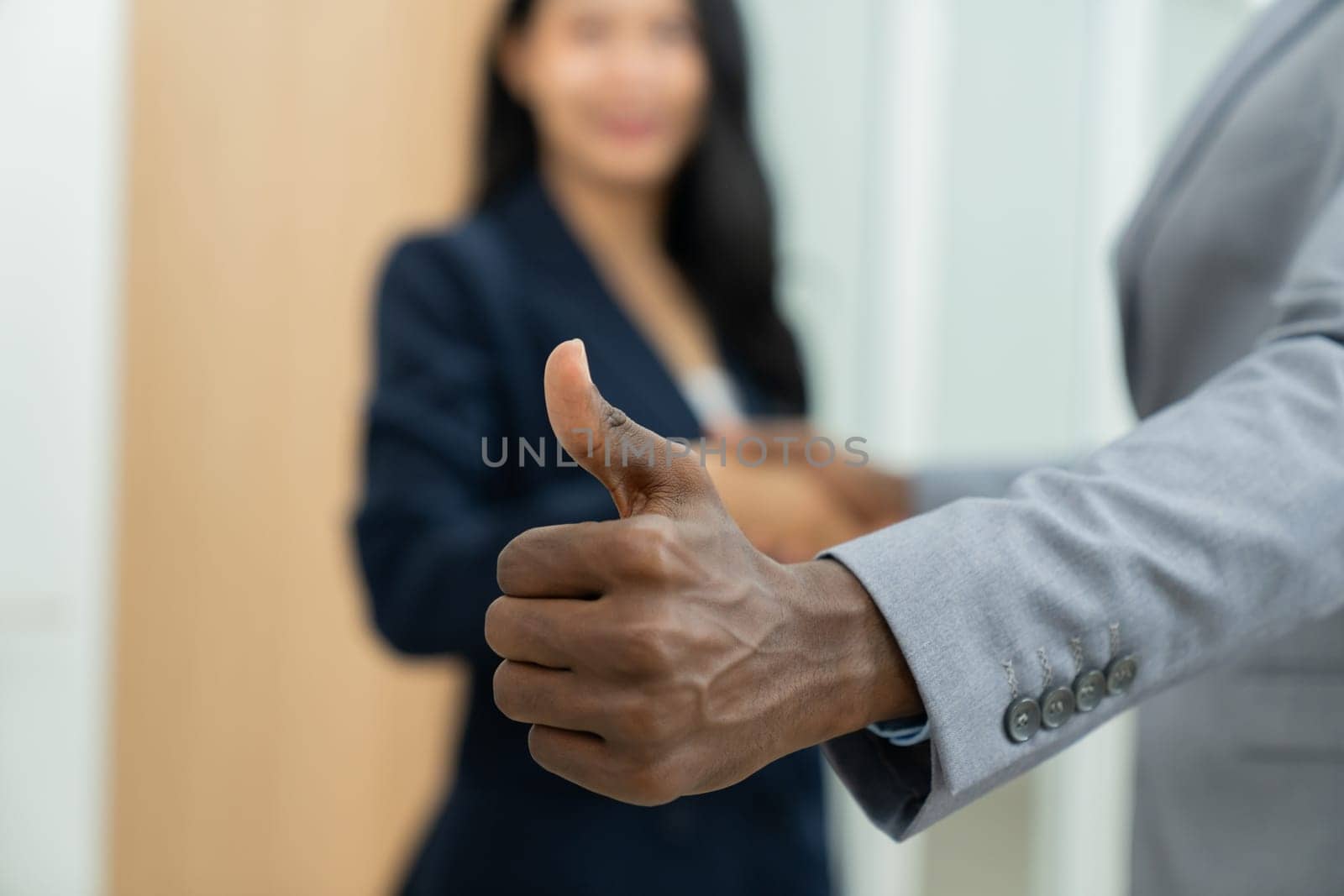 Closeup image of african businessman hand showing thump up to camera while young businesswoman standing behind. Represent recommend, support, good, positive, professional, reliable. Ornamented.