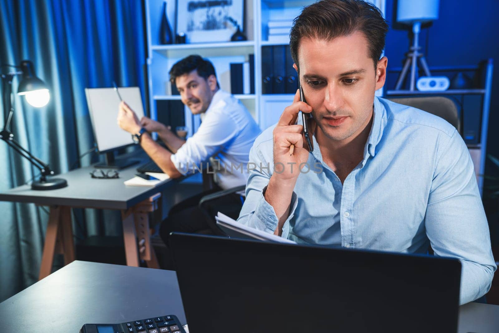 Serious businessman calling on phone with customer to create new project at night time, coworker writing document work sheet on desk. Concept of working on neon blue light modern office. Sellable.