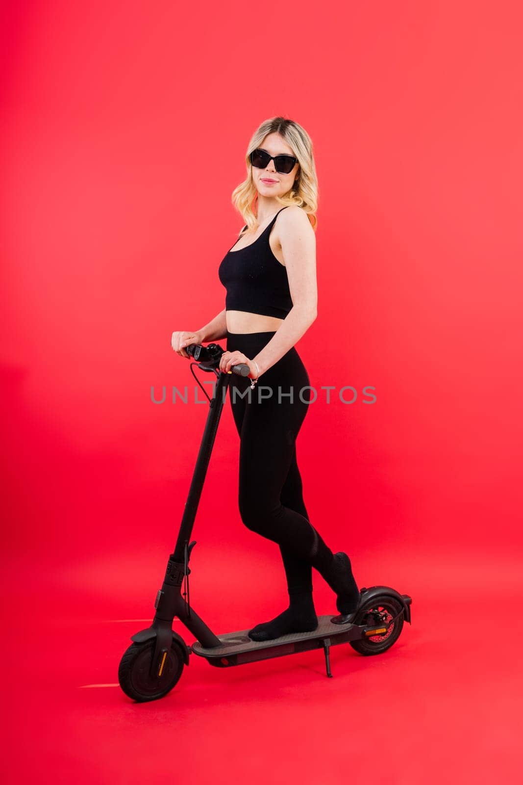 Blonde woman on red white background sitting on electro scooter and resting, copy space by Zelenin