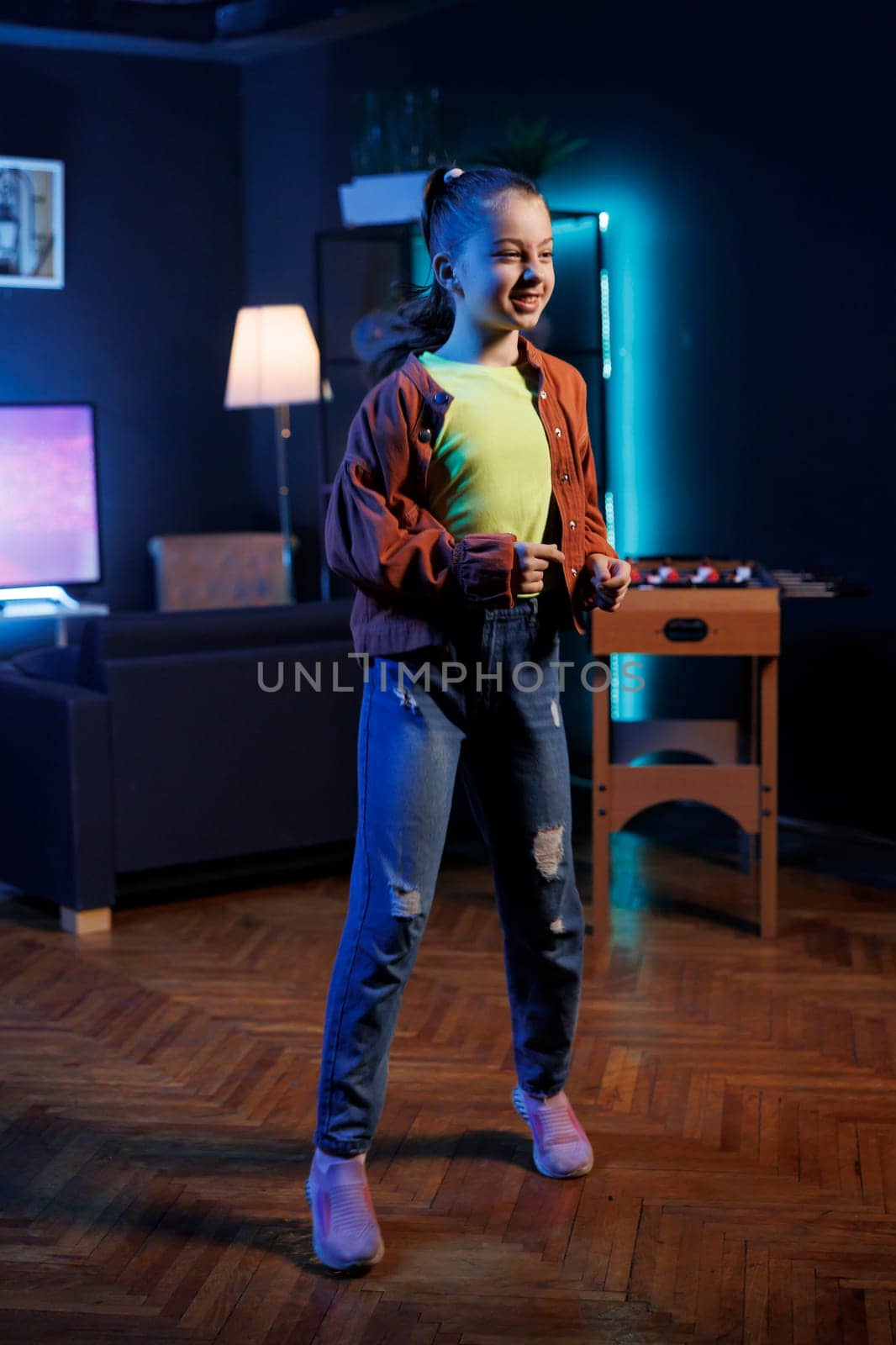 Young girl dancing in dimly lit home studio interior, producing content for online channel. Generation Z youngster doing viral dance choreography in apartment illuminated by RGB lights