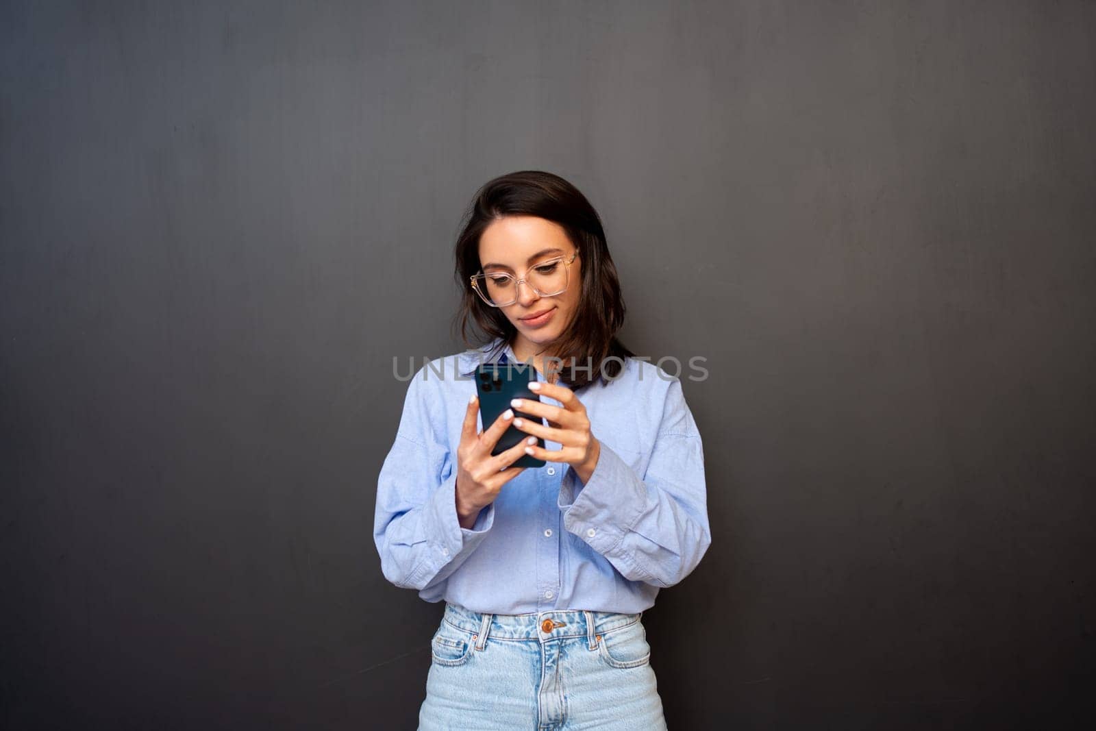 Pensive woman in glasses use smartphone, types text message on mobile phone, enjoys online communication, types feedback, wears denim shirt, isolated on black studio background. Technology concept