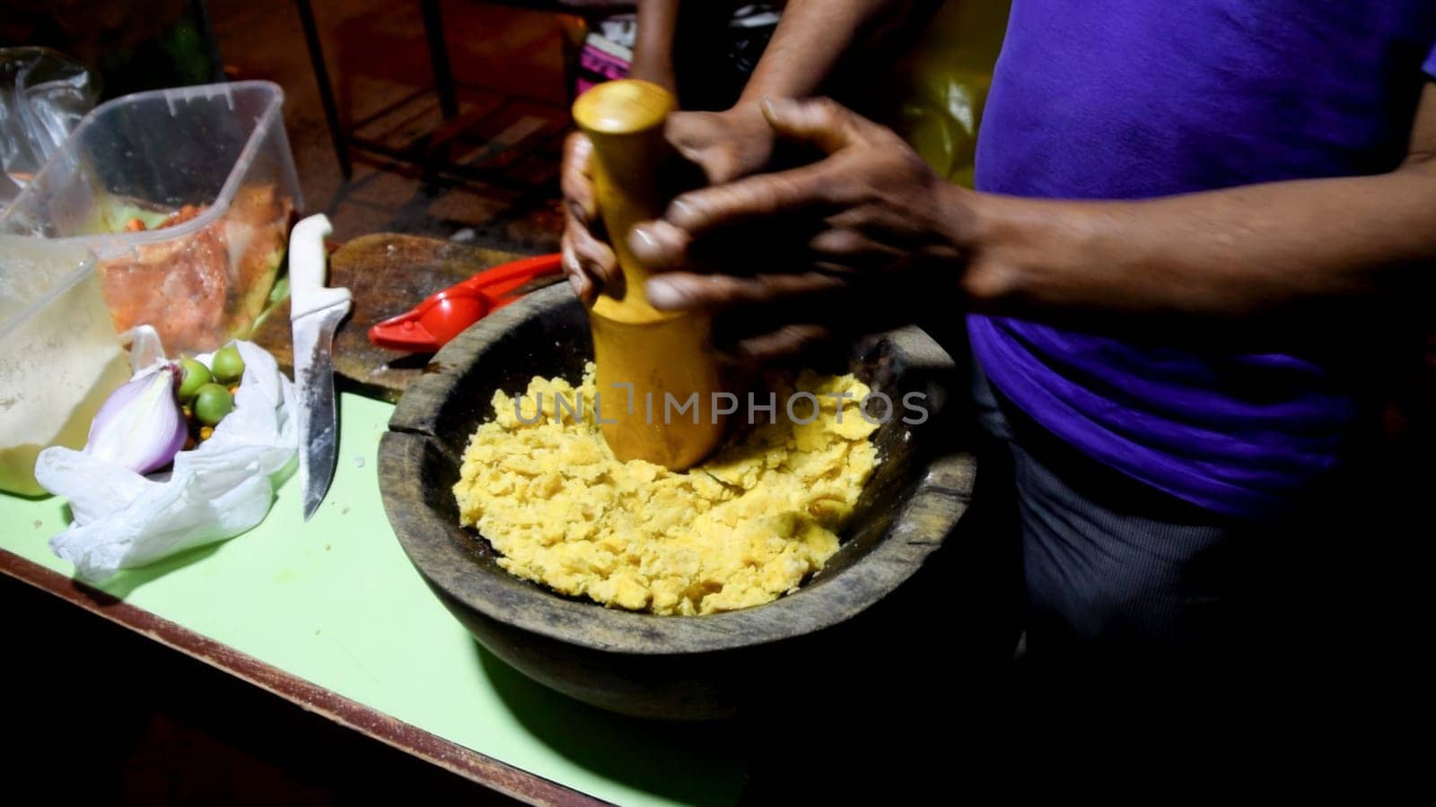 Hands preparing food in a wooden mortar with ingredients nearby on a rustic kitchen table