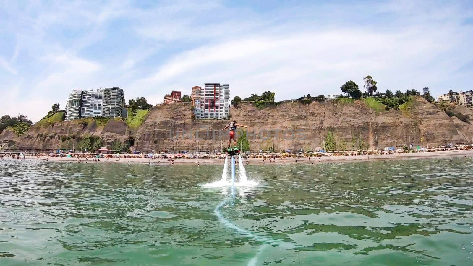 Adventurous individual soars above water using a jetpack, with a scenic city cliffside and beach in the background
