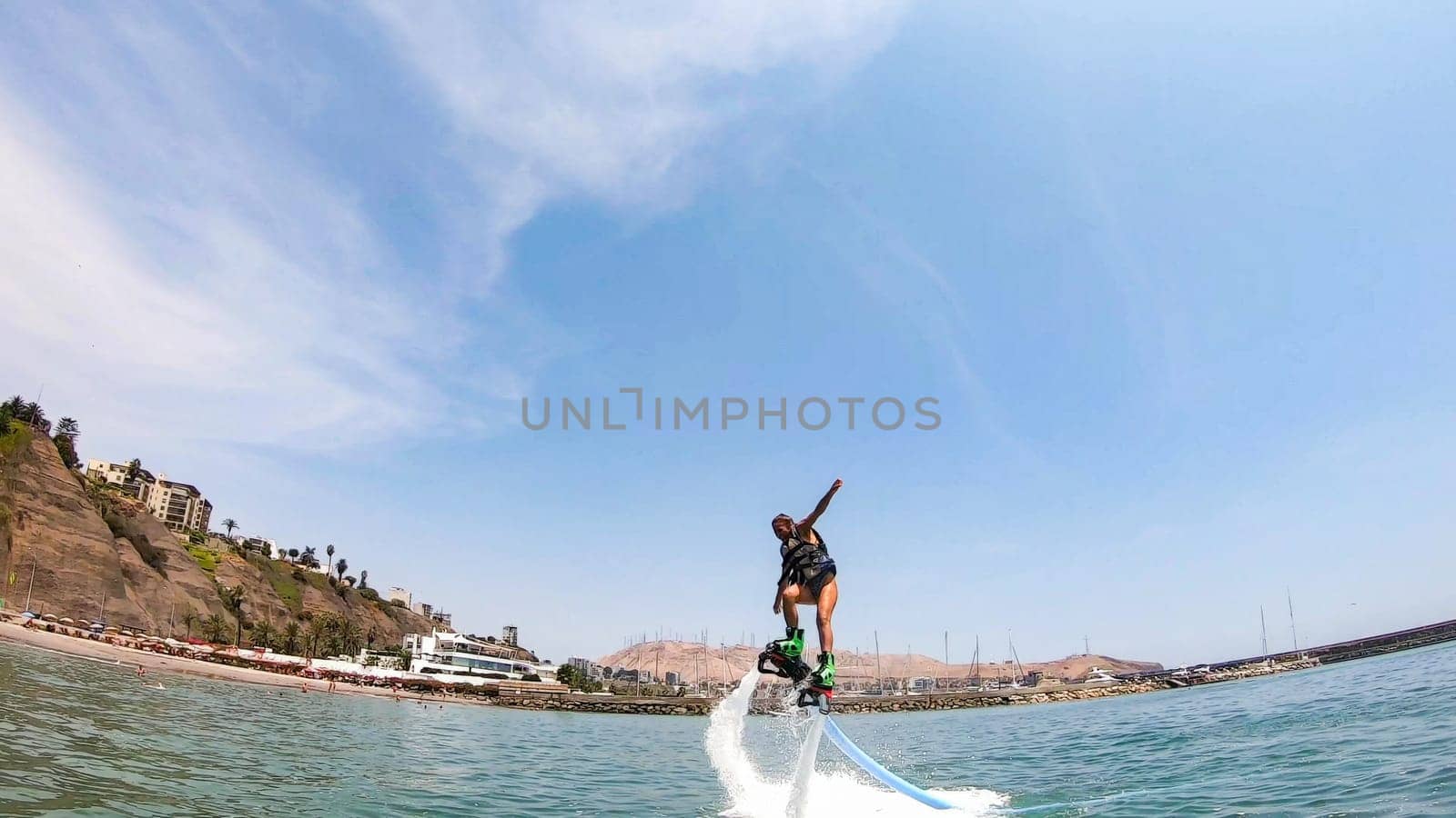 Exhilarating shot of a person flyboarding above the sea with a coastal cityscape backdrop