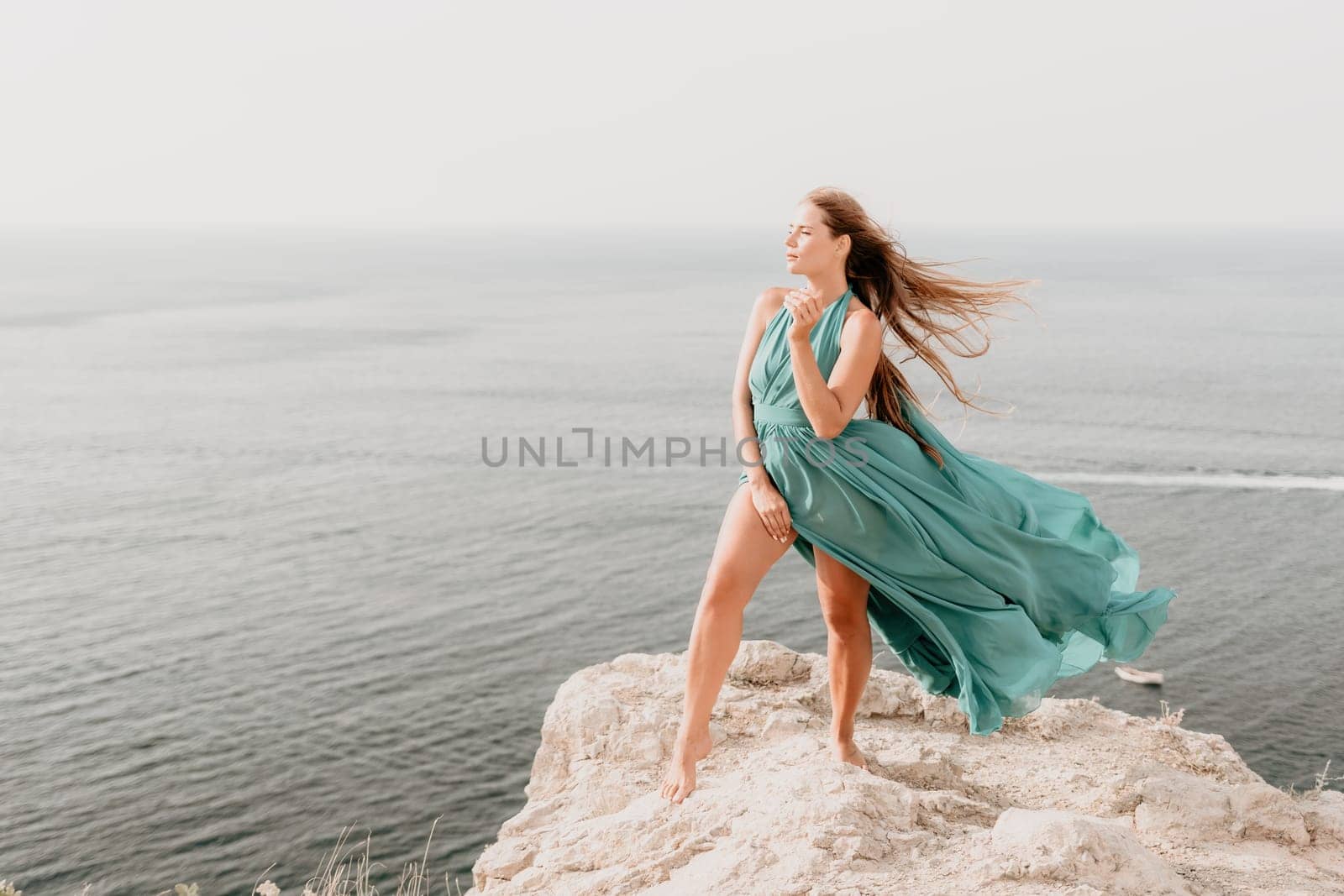 Side view a Young beautiful sensual woman in a mint long dress posing on a volcanic rock high above the sea during sunset. Girl on the nature on overcast sky background. Fashion photo