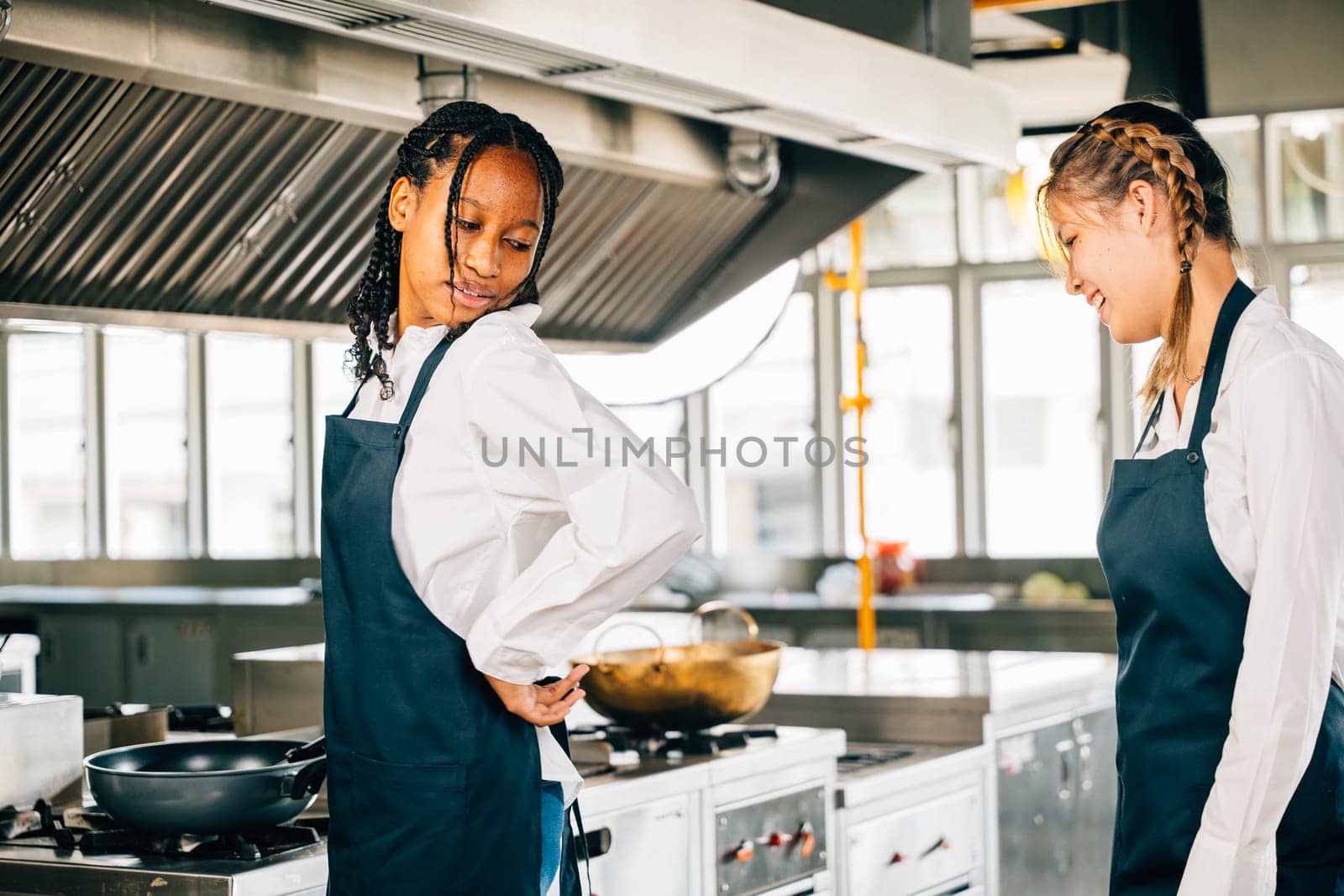Female chef aids friend tie apron in a restaurant kitchen. Two adults in uniform prepare for food education and service in a professional setup. by Sorapop