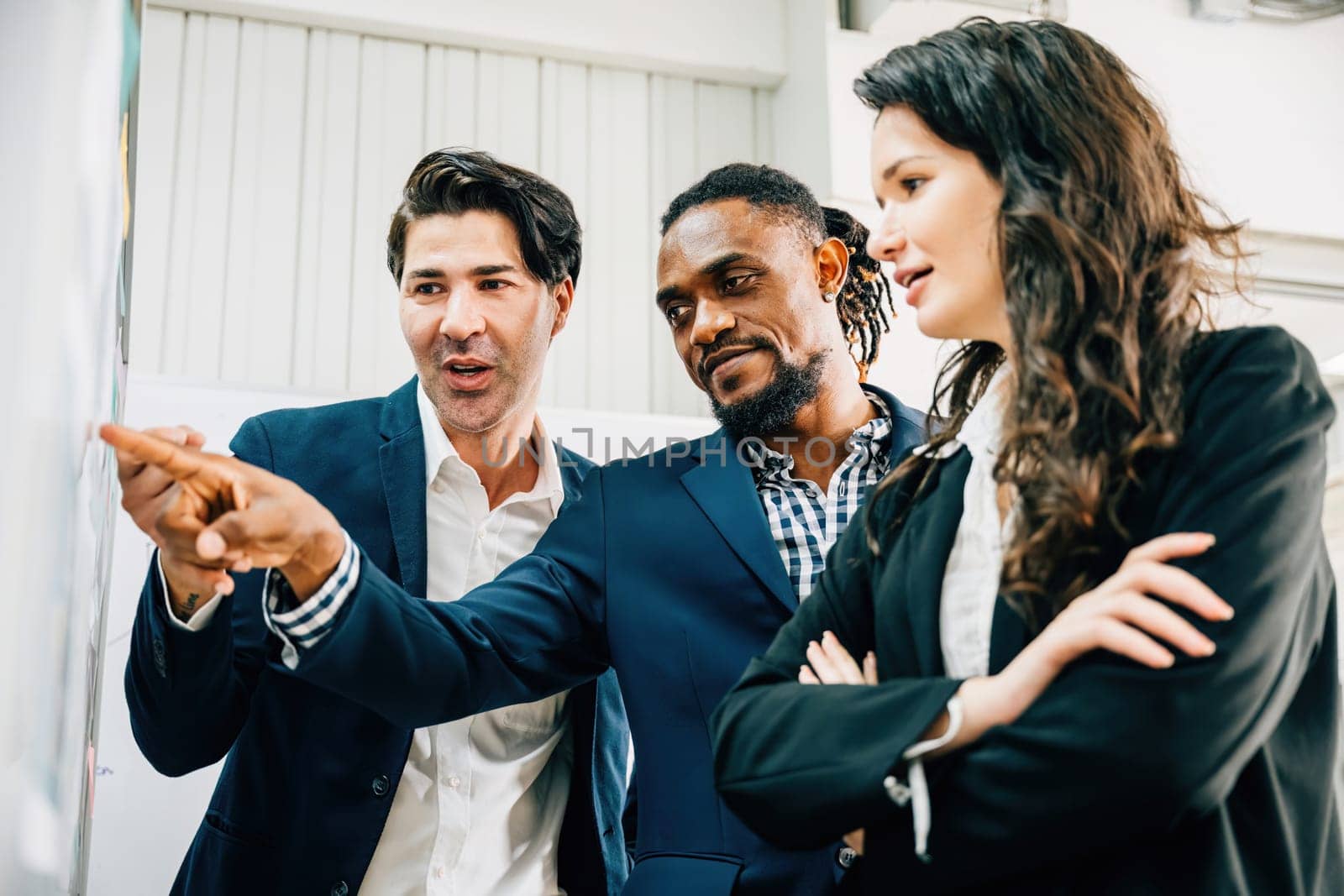 In an office setting, business professionals engage in brainstorming with post-it notes on a glass wall. Their diversity underlines teamwork, leadership, and fruitful business discussions. by Sorapop