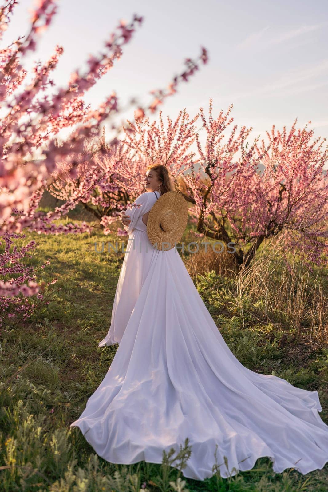 Woman blooming peach orchard. Against the backdrop of a picturesque peach orchard, a woman in a long white dress and hat enjoys a peaceful walk in the park, surrounded by the beauty of nature