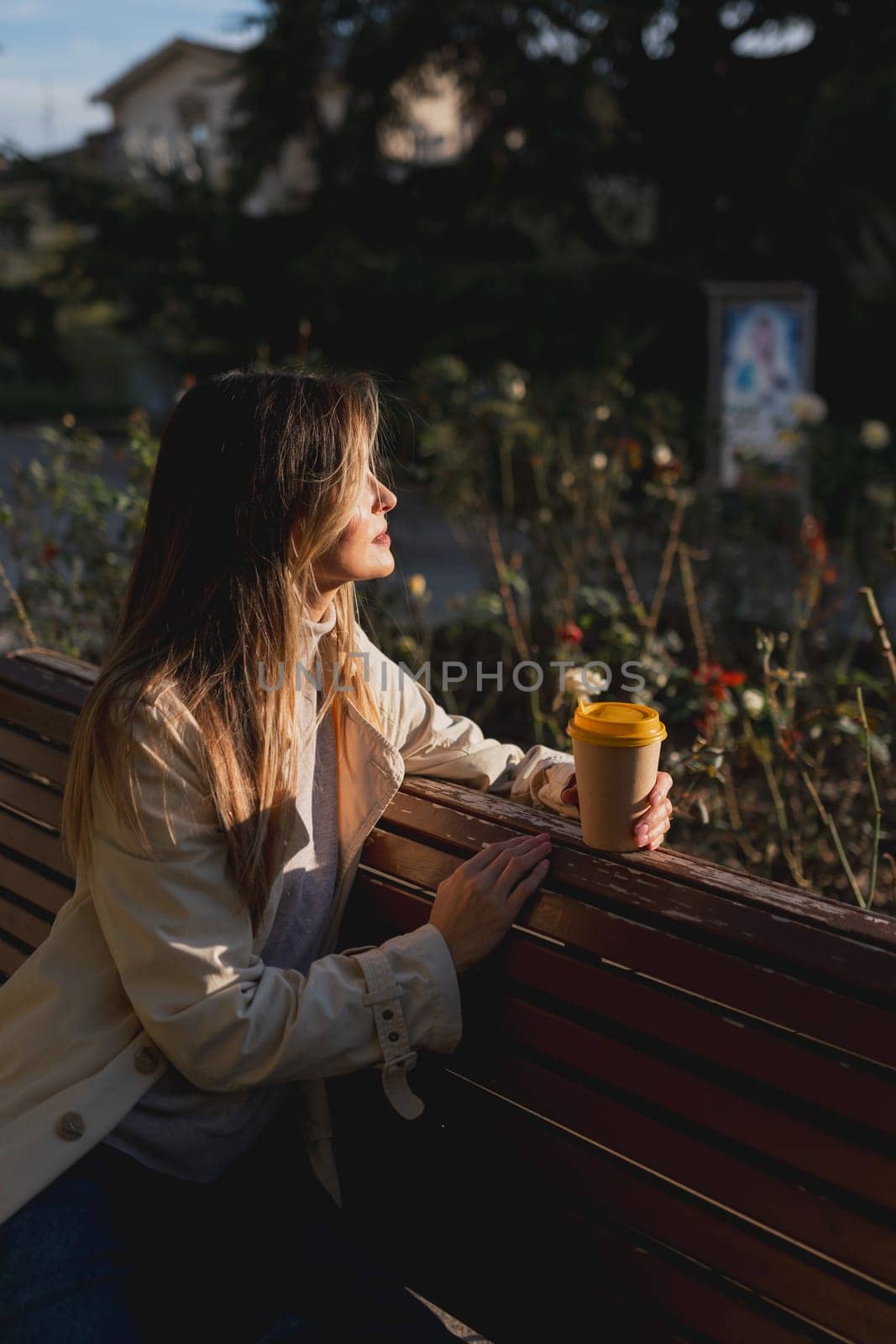 Woman drinks from cup on wooden bench. She is wearing a white shirt enjoying her beverage. The bench is located in a park setting, with trees in the background. by Matiunina