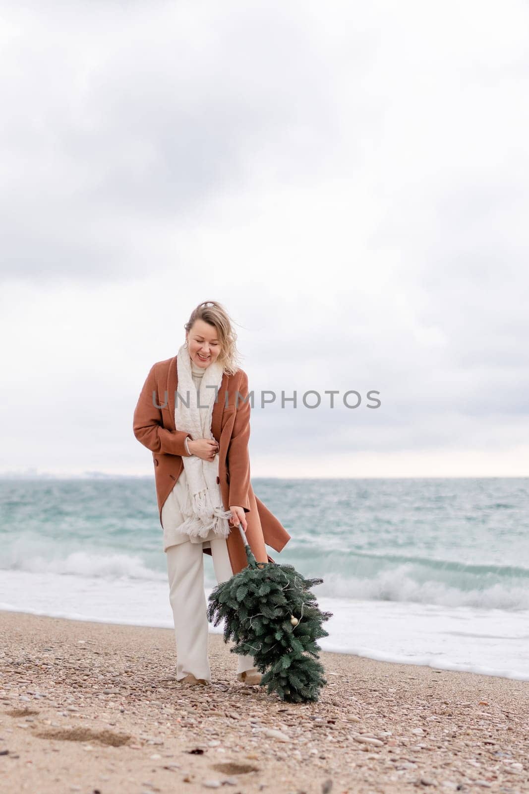 Blond woman Christmas sea. Christmas portrait of a happy woman walking along the beach and holding a Christmas tree in her hands. She is wearing a brown coat and a white suit